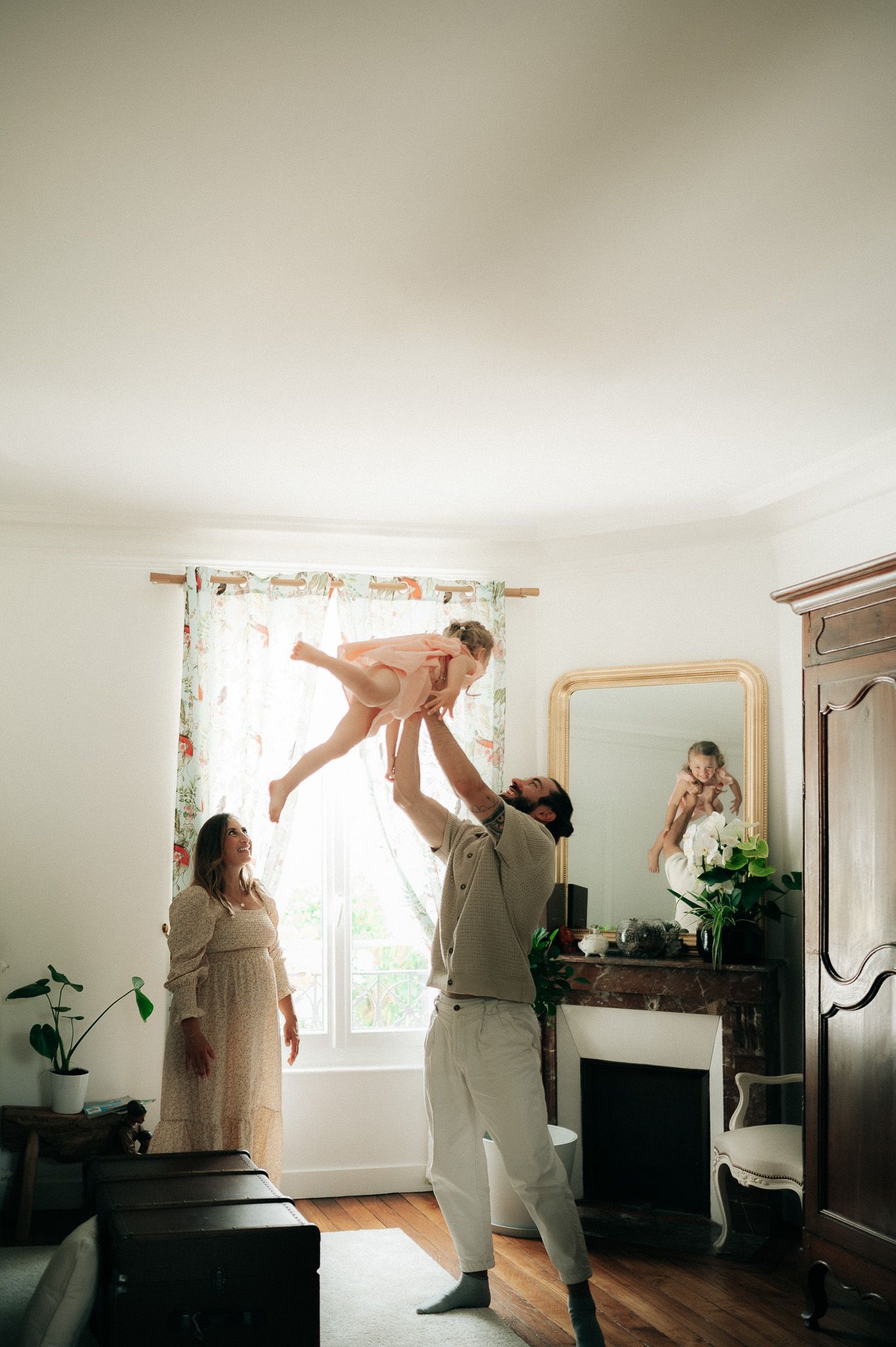 Father tossing young daughter in air while mother watches in bright sunlit family home.