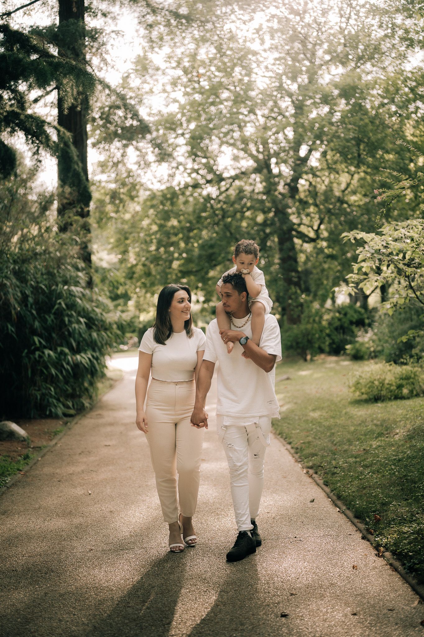 Family of three walking hand in hand on a garden path, dressed in coordinating white outfits, with child on father's shoulders.