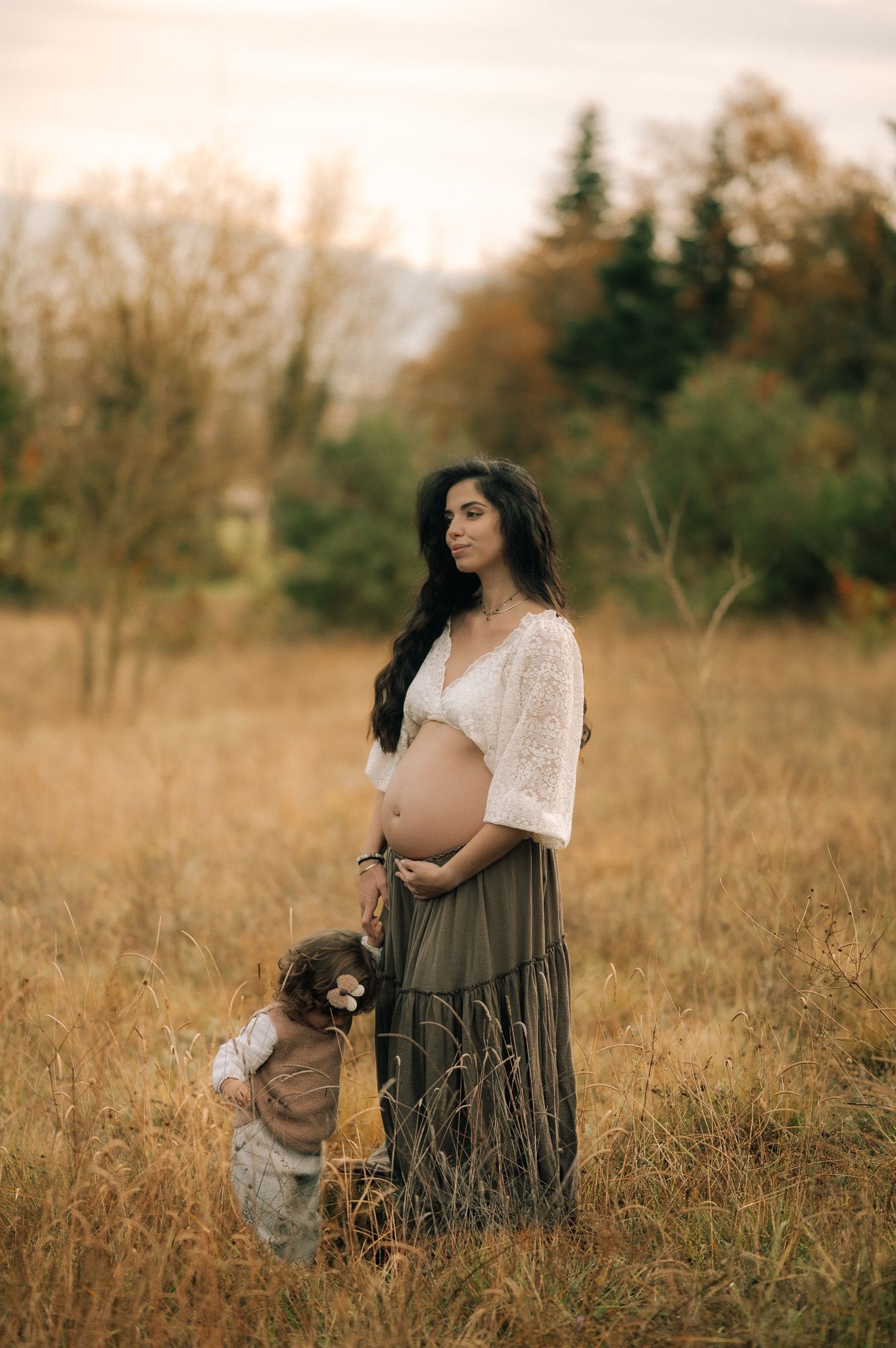 Pregnant woman in white lace top and olive skirt stands in golden field with toddler child during autumn maternity photoshoot.