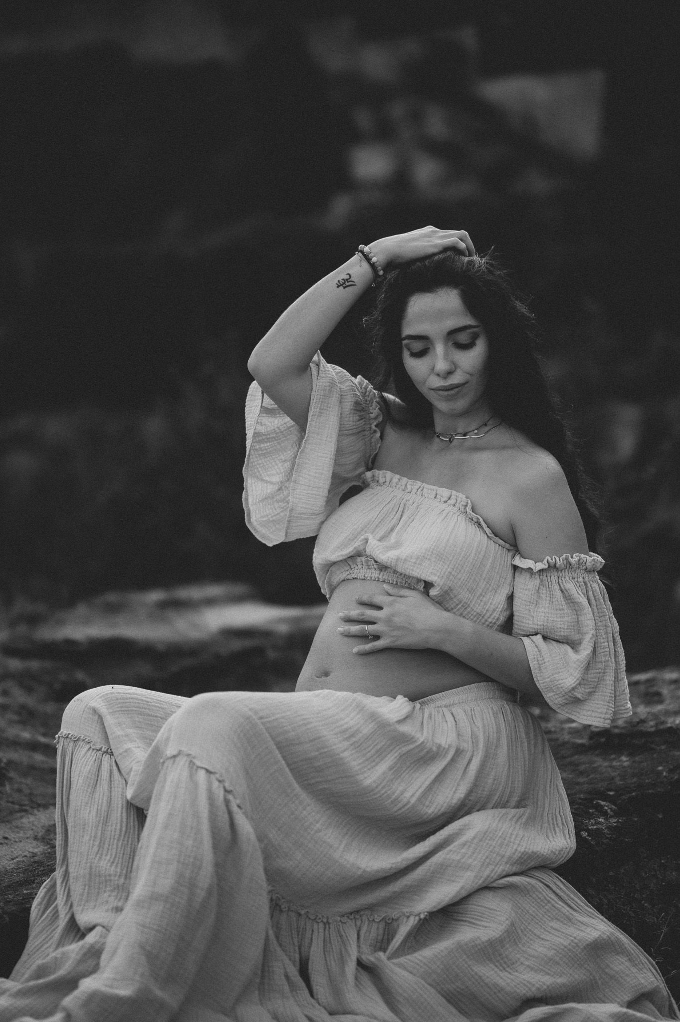 Black and white maternity photo of a woman in an off-shoulder dress sitting outdoors, cradling her baby bump.