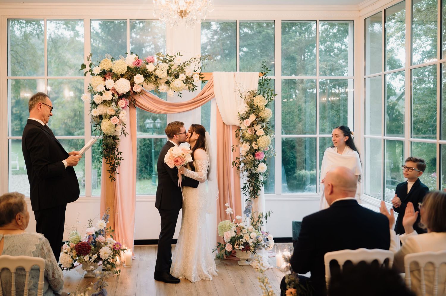 Bride and groom share their first kiss under a floral arch while guests applaud the ceremony.
