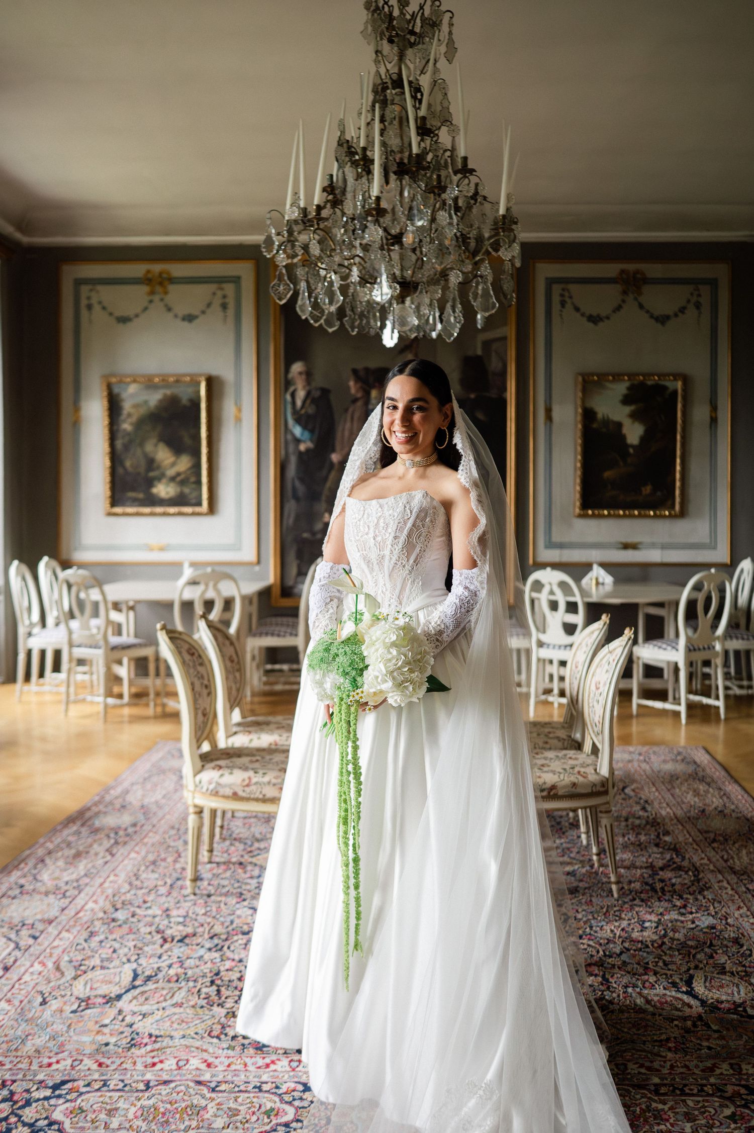 Smiling bride in white gown and veil holds cascading bouquet under crystal chandelier in elegant hall.