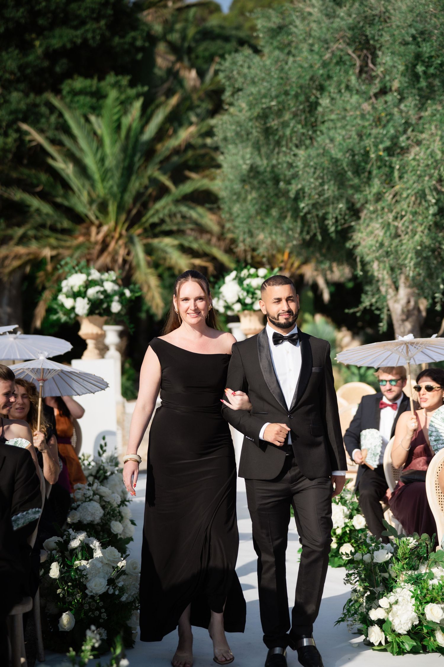 Groom in black tuxedo walks down the aisle with a woman in a black gown at an outdoor wedding.