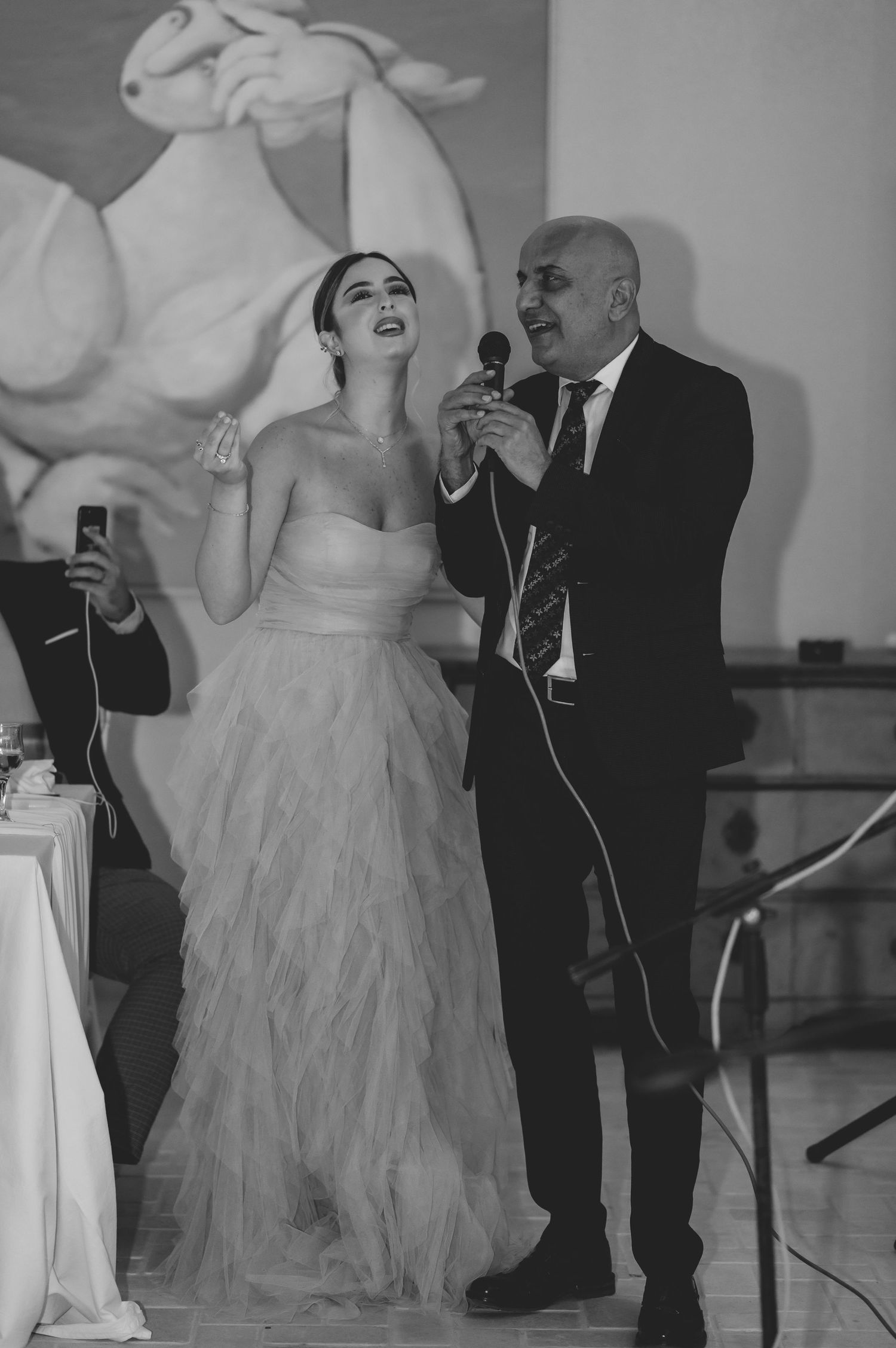 Black and white photo of bride in strapless gown singing with older man holding microphone at reception.