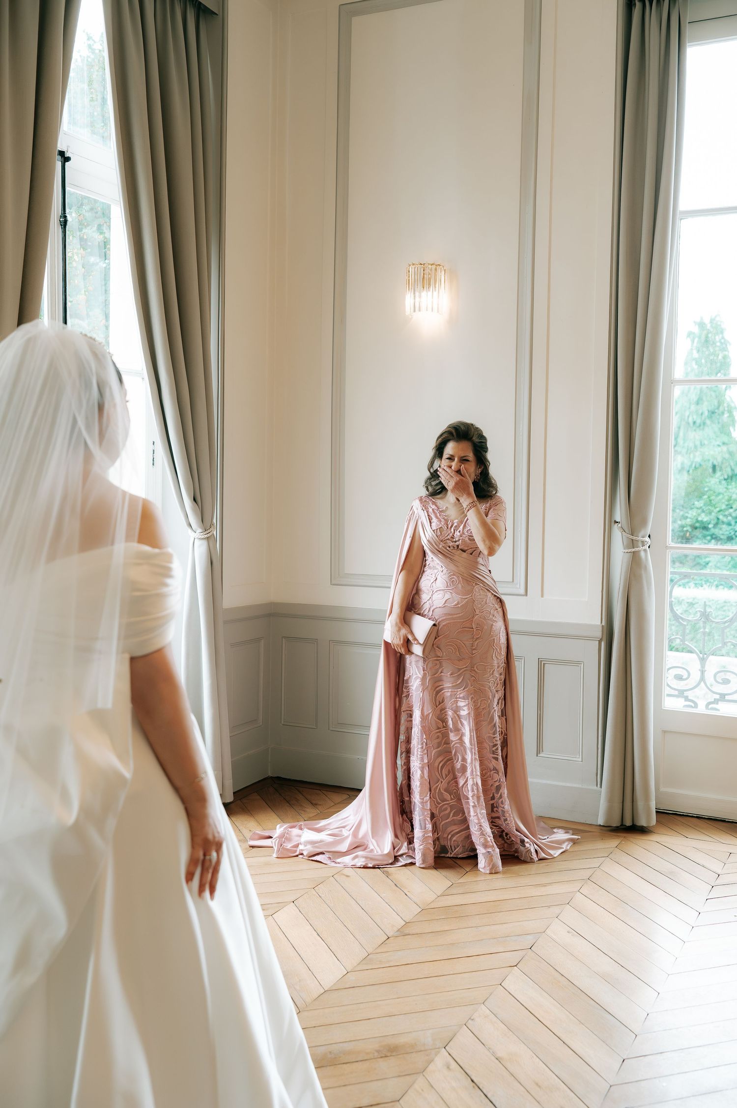 Emotional mother in pink gown reacts to seeing bride in white wedding dress during first look.