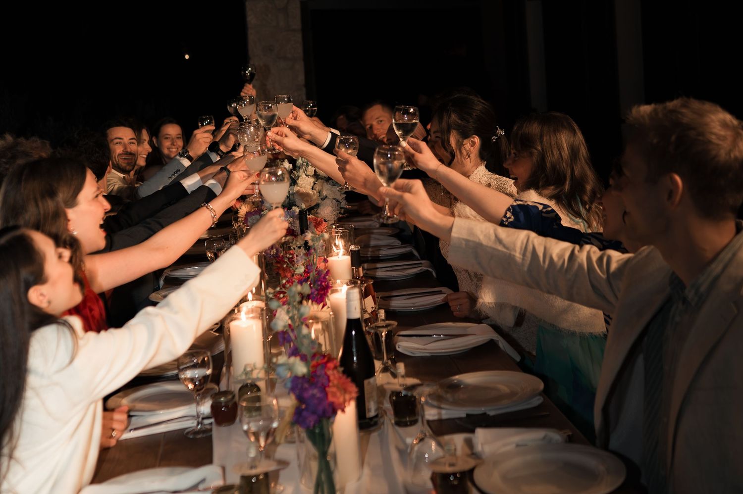 Wedding guests raise glasses in a joyful toast over a flower-adorned candlelit reception dinner table.