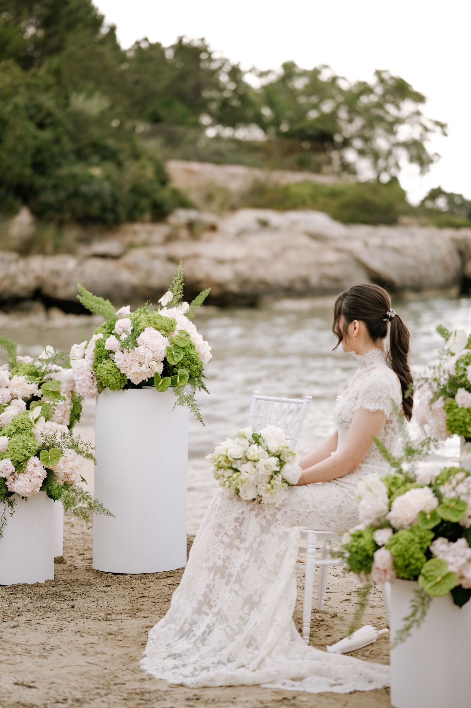 Bride in lace gown sits amid white floral pedestals during seaside wedding ceremony.