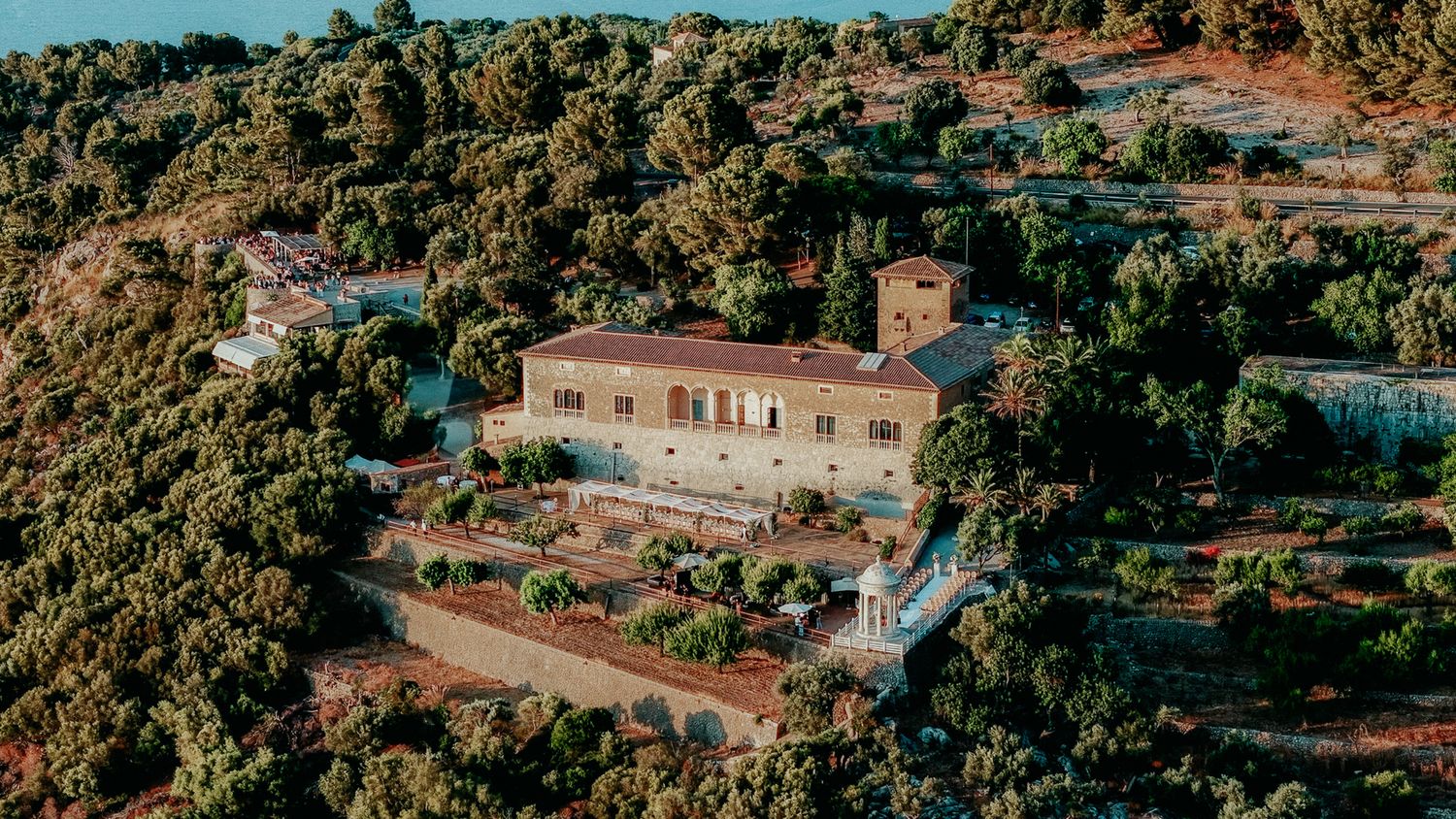 Aerial view of a large stone manor house surrounded by lush Mediterranean hillside vegetation at golden hour.