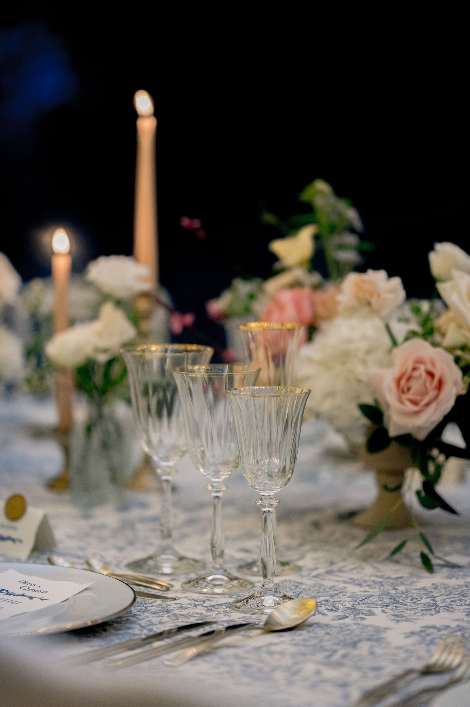 Close-up of crystal champagne flutes and pink roses on a blue toile wedding reception table with candlelight.