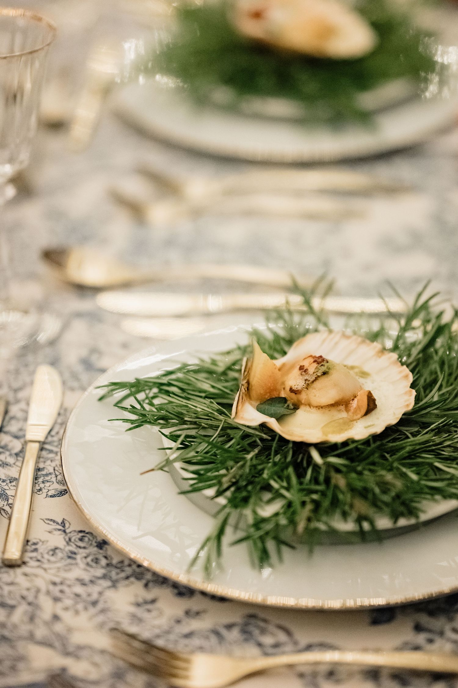 Elegant wedding place setting with a scallop shell appetizer on fresh herbs atop a gold-rimmed plate.