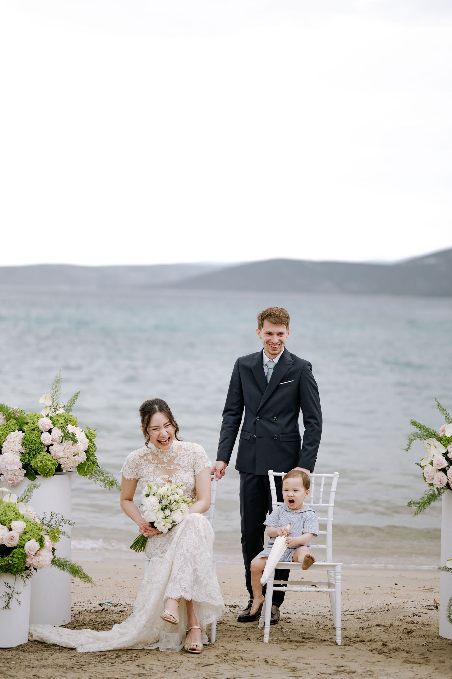 Smiling bride and groom stand with young child in white chair at elegant beach wedding ceremony.
