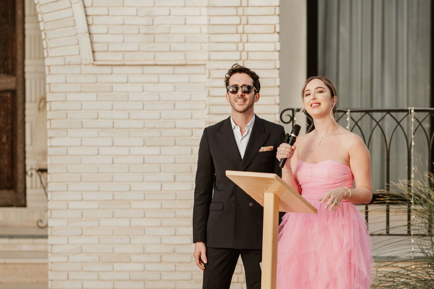 Man in black suit and woman in pink strapless gown speak at podium during outdoor wedding ceremony.