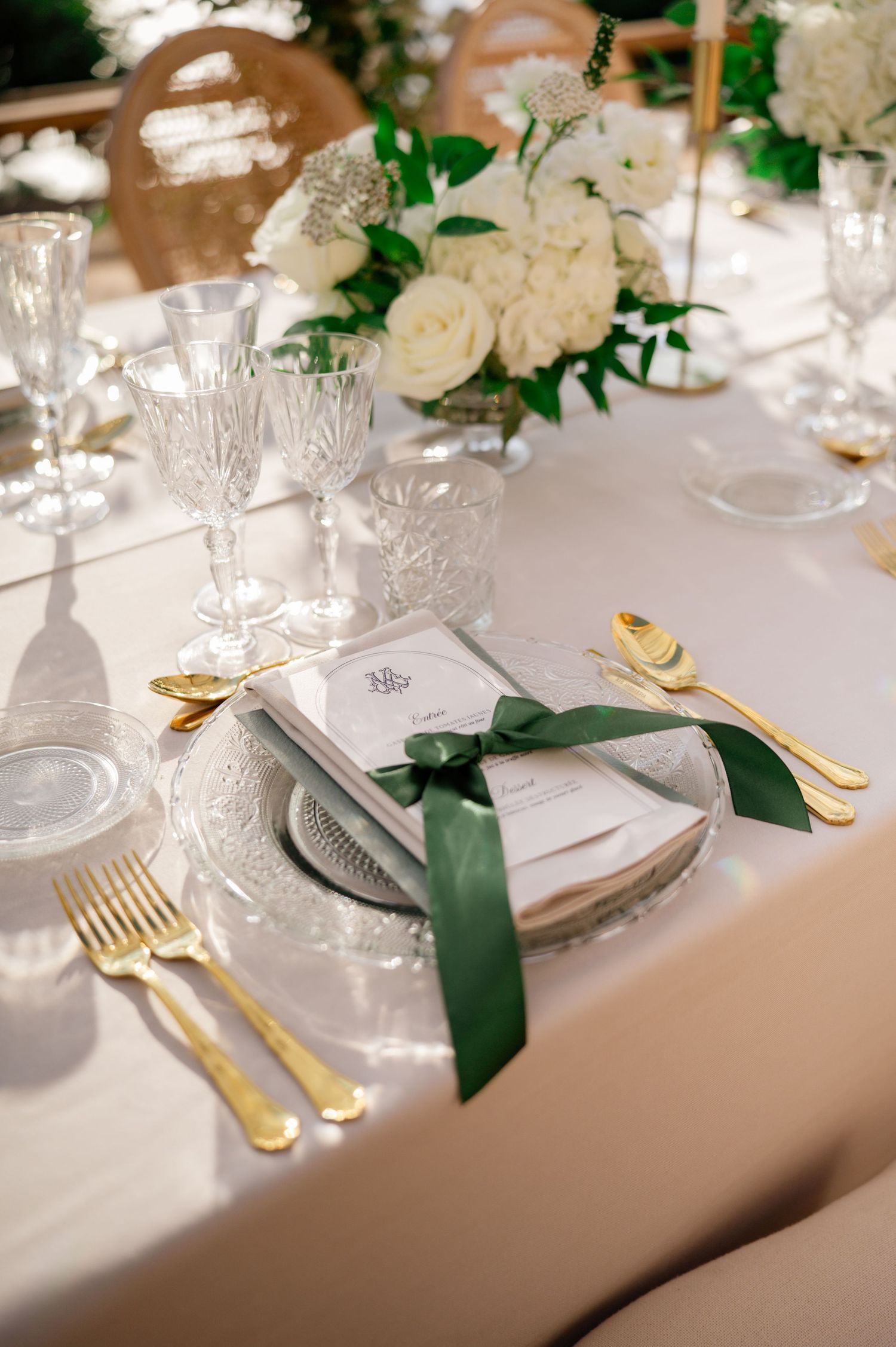 Elegant wedding place setting with gold cutlery, crystal glasses, green ribbon-tied menu card, and white floral centerpiece.