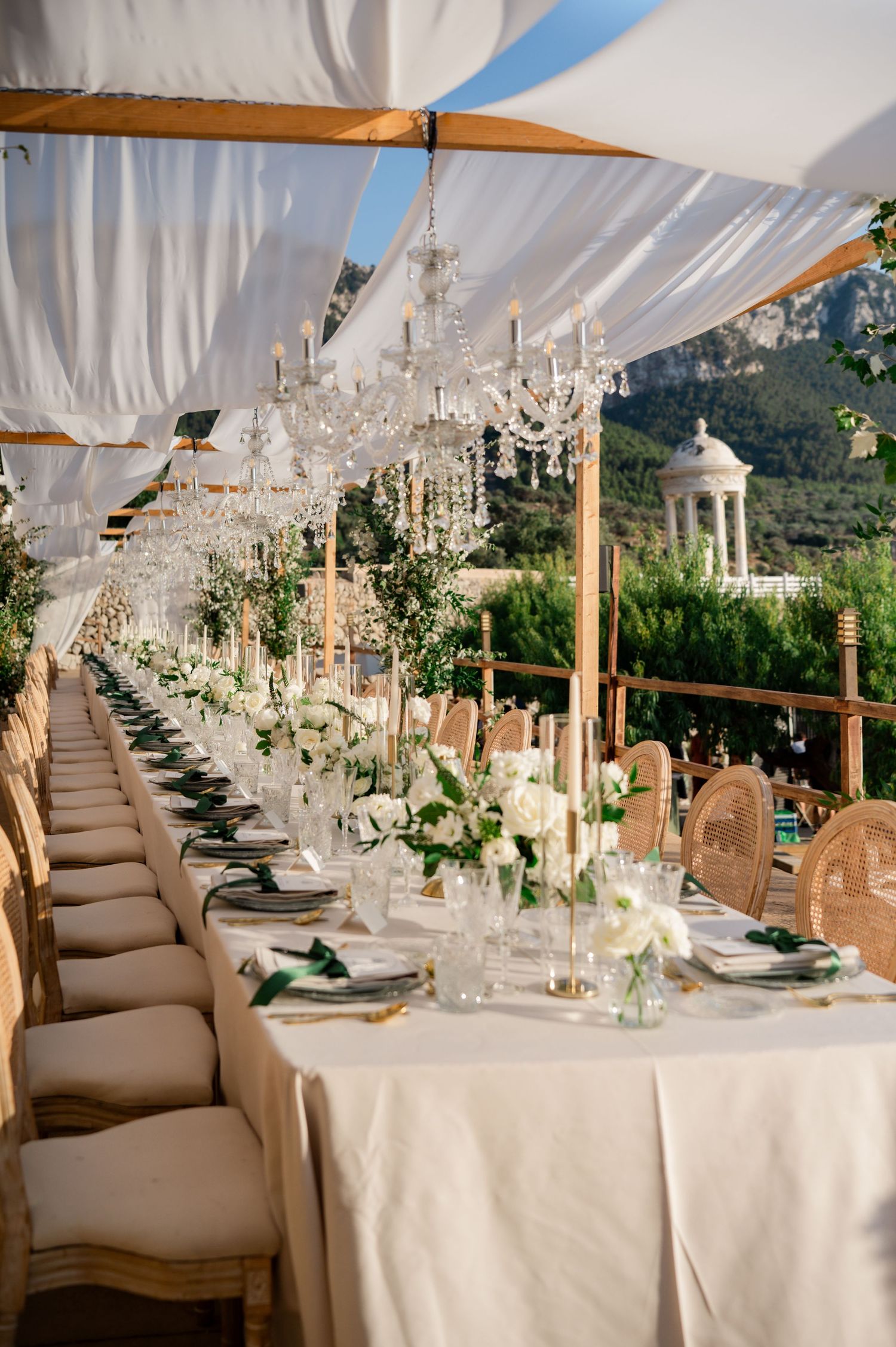 Elegant outdoor wedding banquet with long table, crystal chandeliers, white florals, wicker chairs, and mountain backdrop.