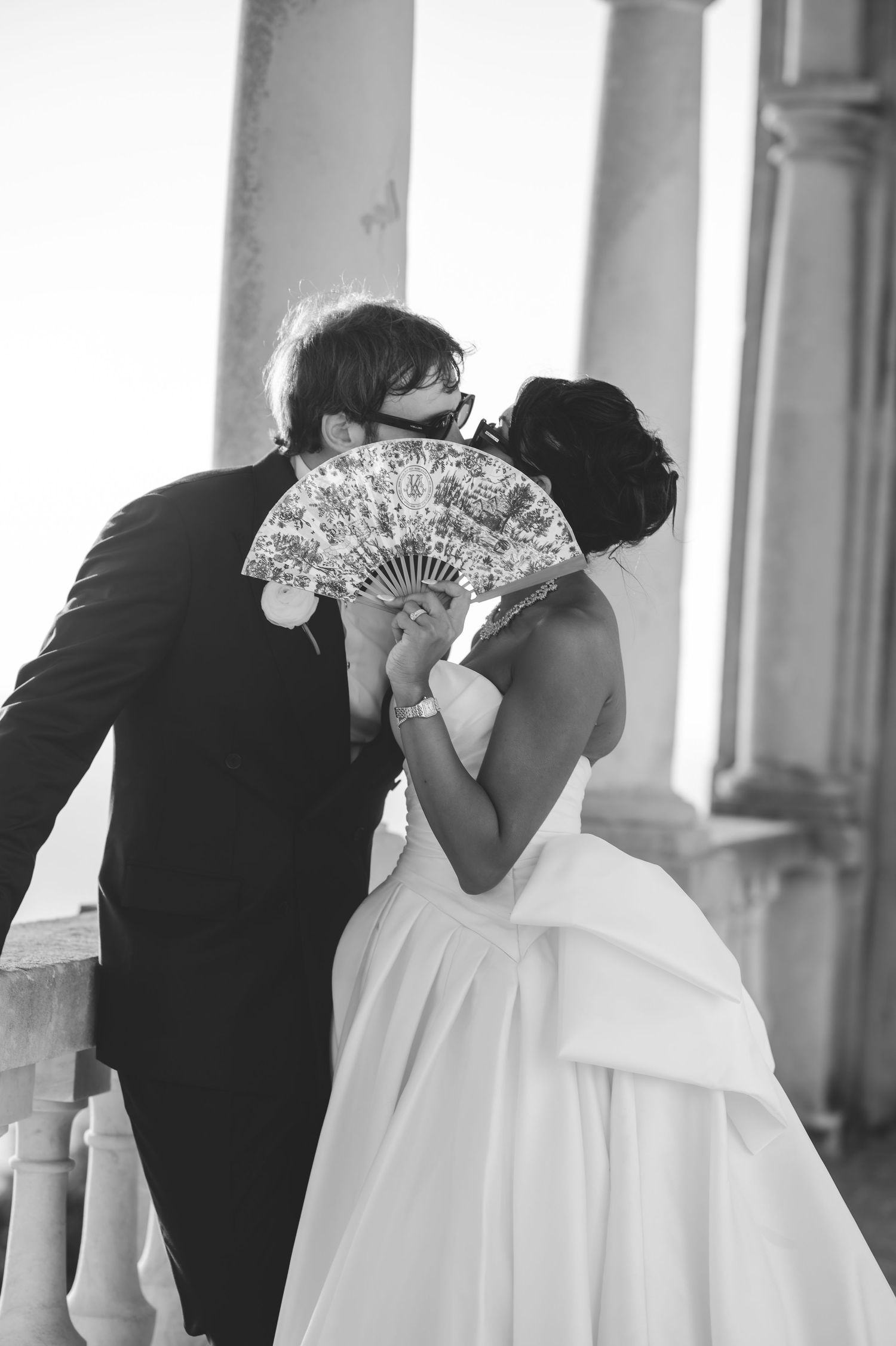 Black and white photo of newlyweds sharing a kiss hidden behind a decorative hand fan.