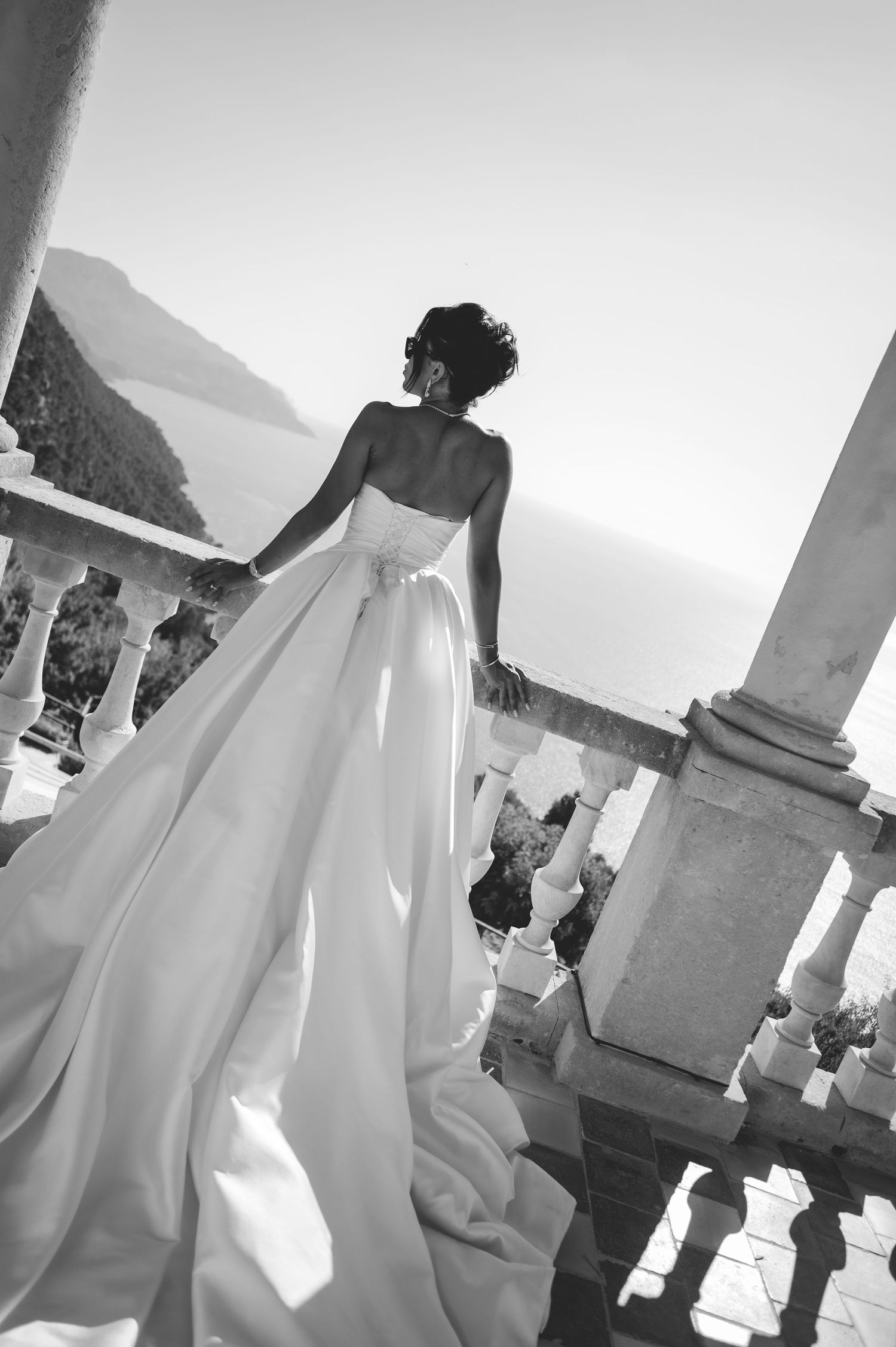 Bride in strapless white ball gown with flowing train stands on marble balcony overlooking sea.