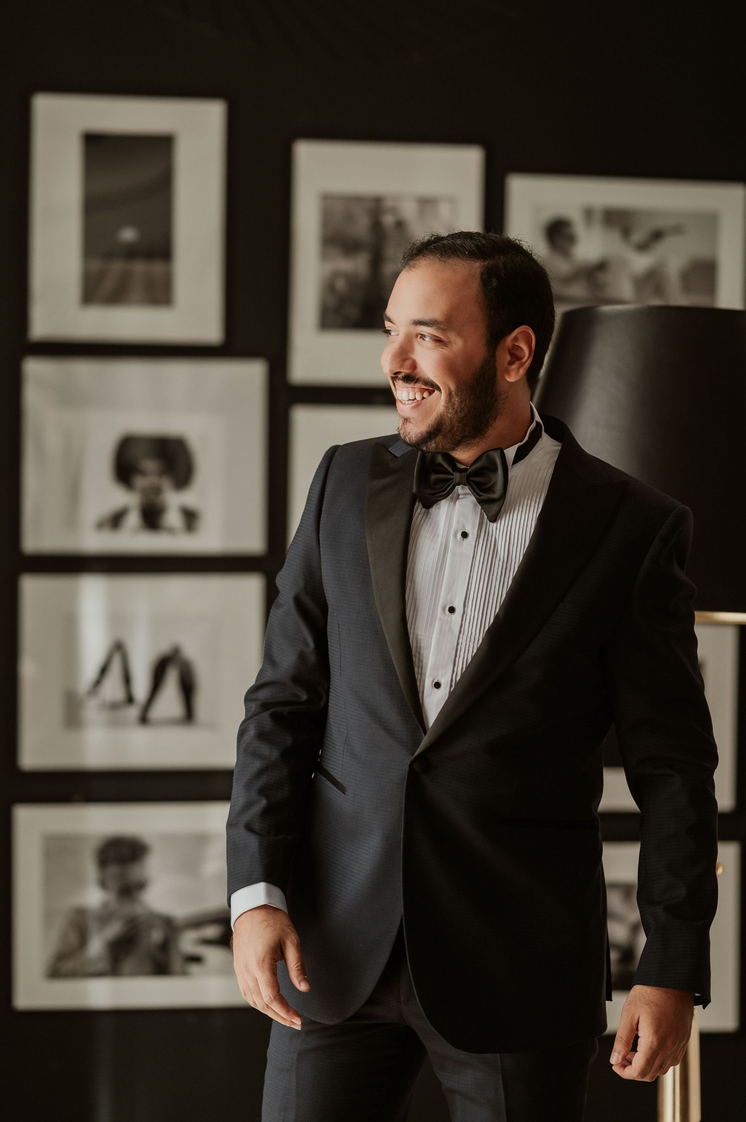 Smiling groom in black tuxedo with bow tie standing in front of black and white framed photos.