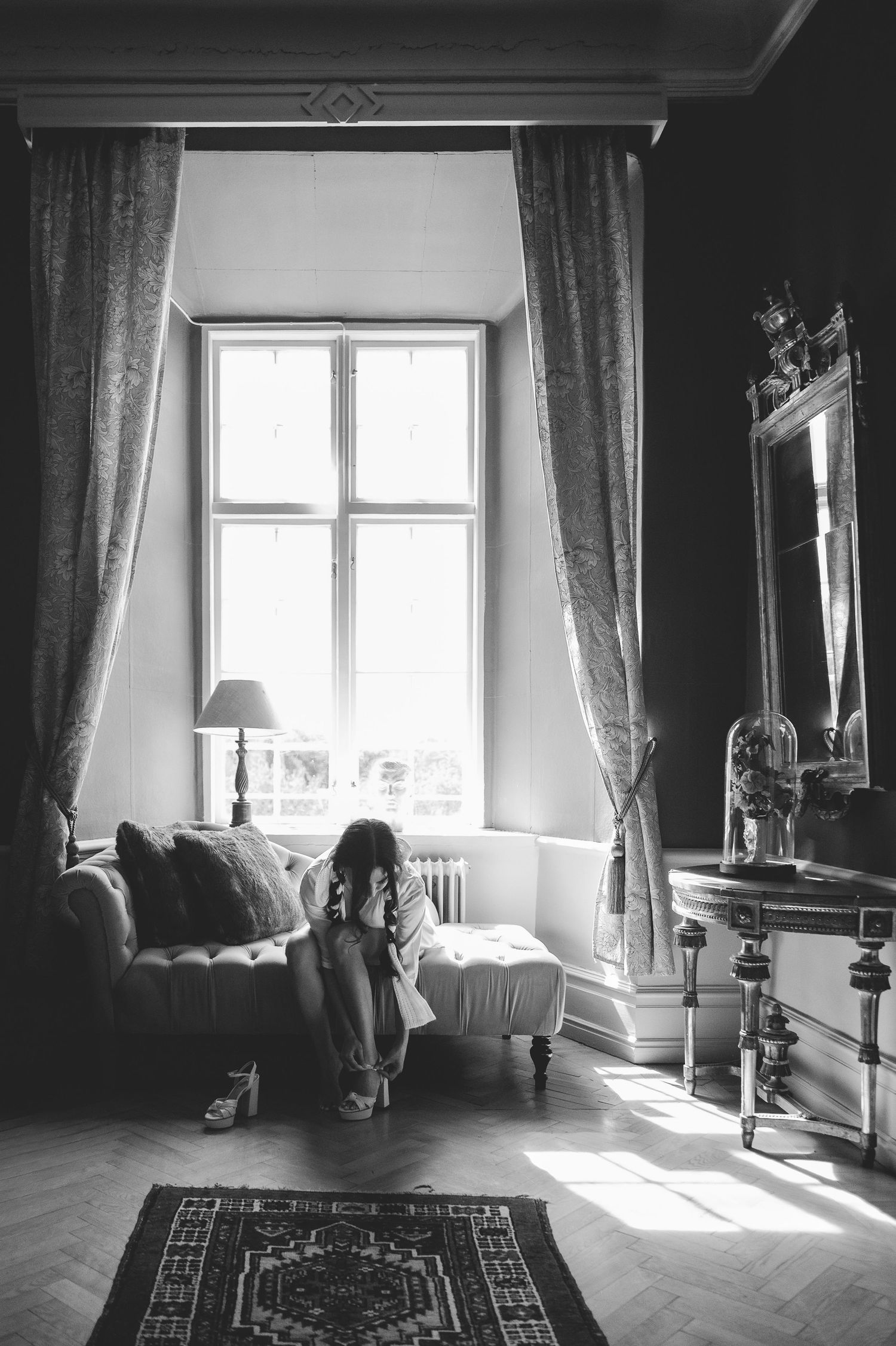 Black and white photo of bride preparing alone by sunlit window with ornate mirror in elegant room.