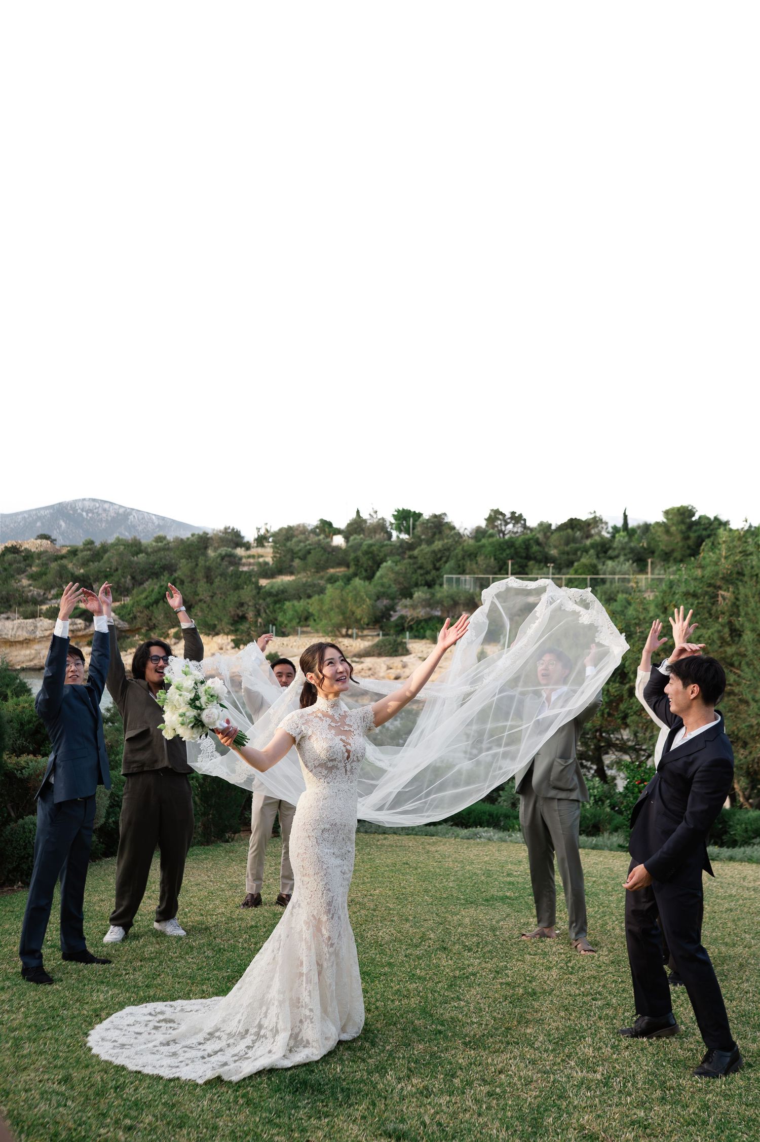 Bride tossing veil with wedding party celebrating outdoors in a scenic garden setting.