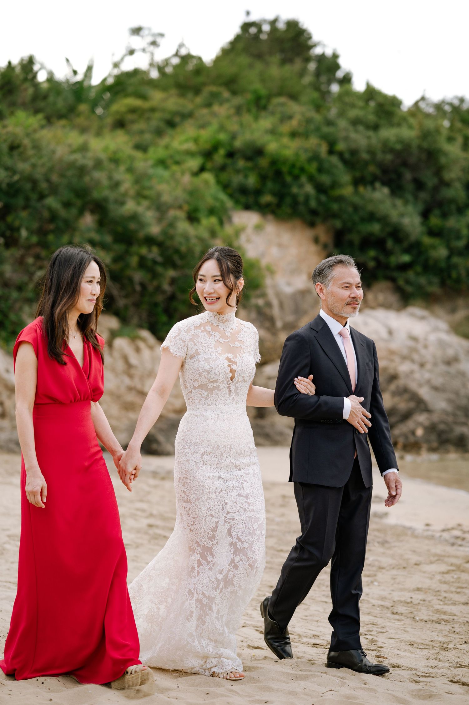 Bride in lace gown walks on sandy beach with parents during wedding processional.