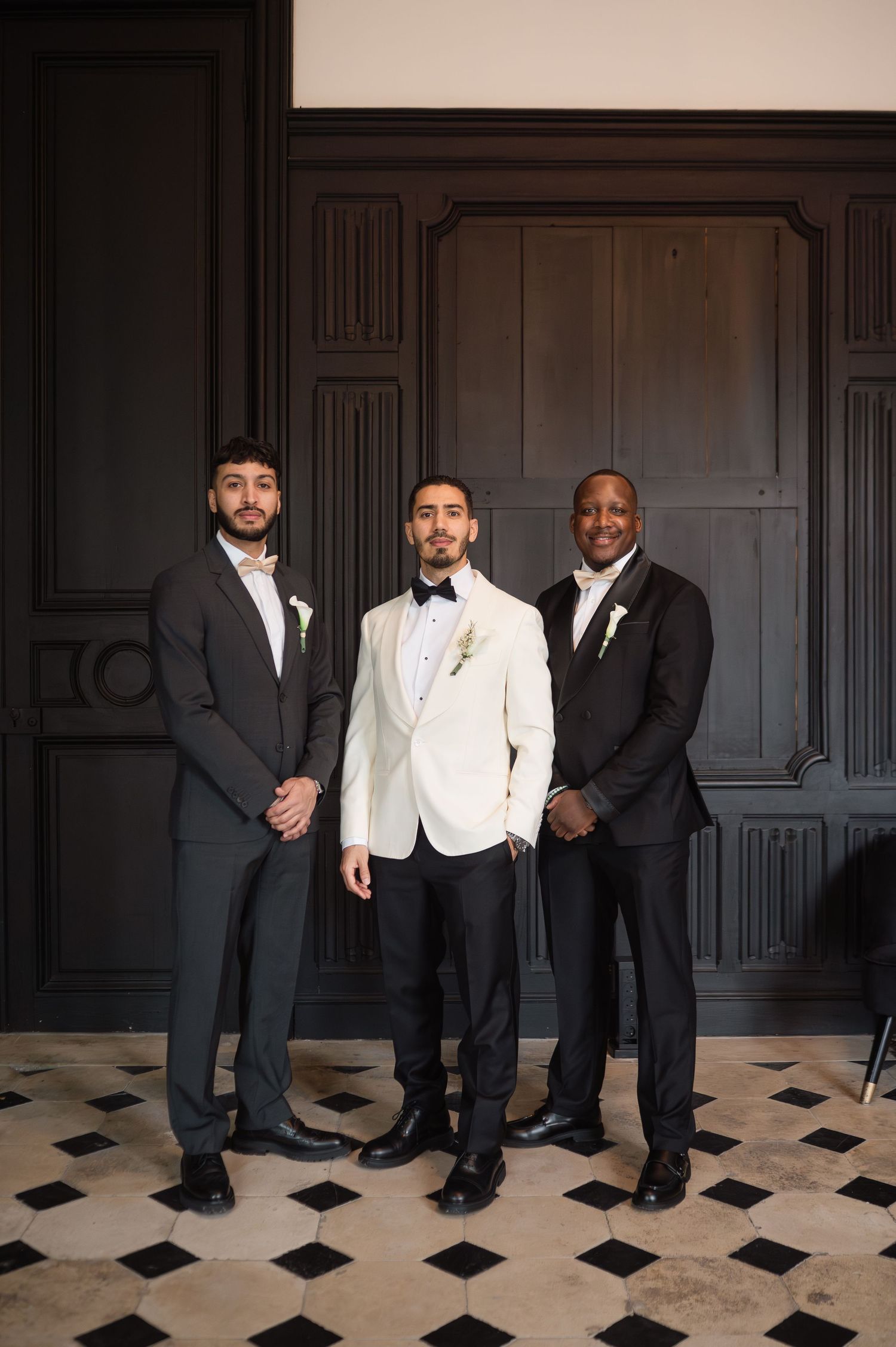 Groom in white dinner jacket poses with two groomsmen against dark wood paneling.
