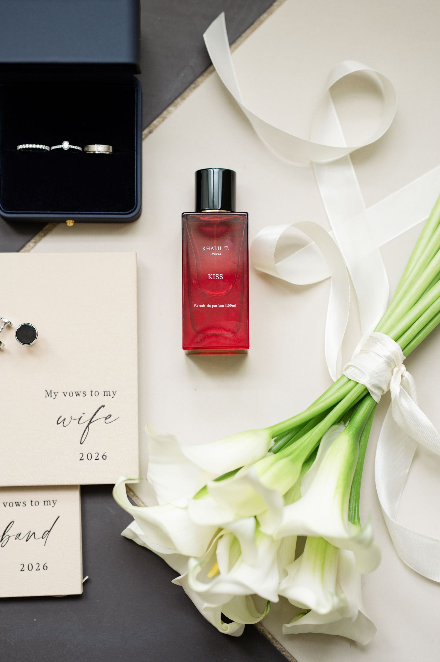 Wedding flat lay with rings, vow books, KISS perfume, calla lily bouquet, and ribbon.