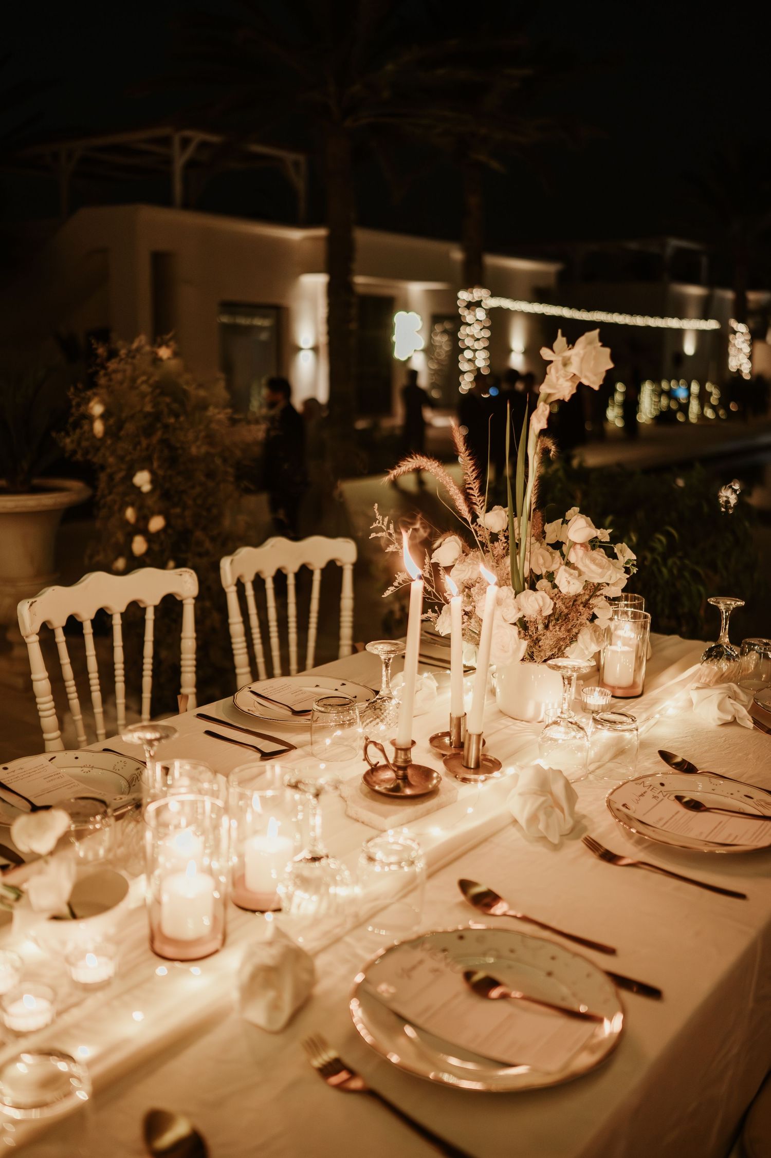 Romantic outdoor wedding reception table with candles, white flowers, and golden cutlery at night.