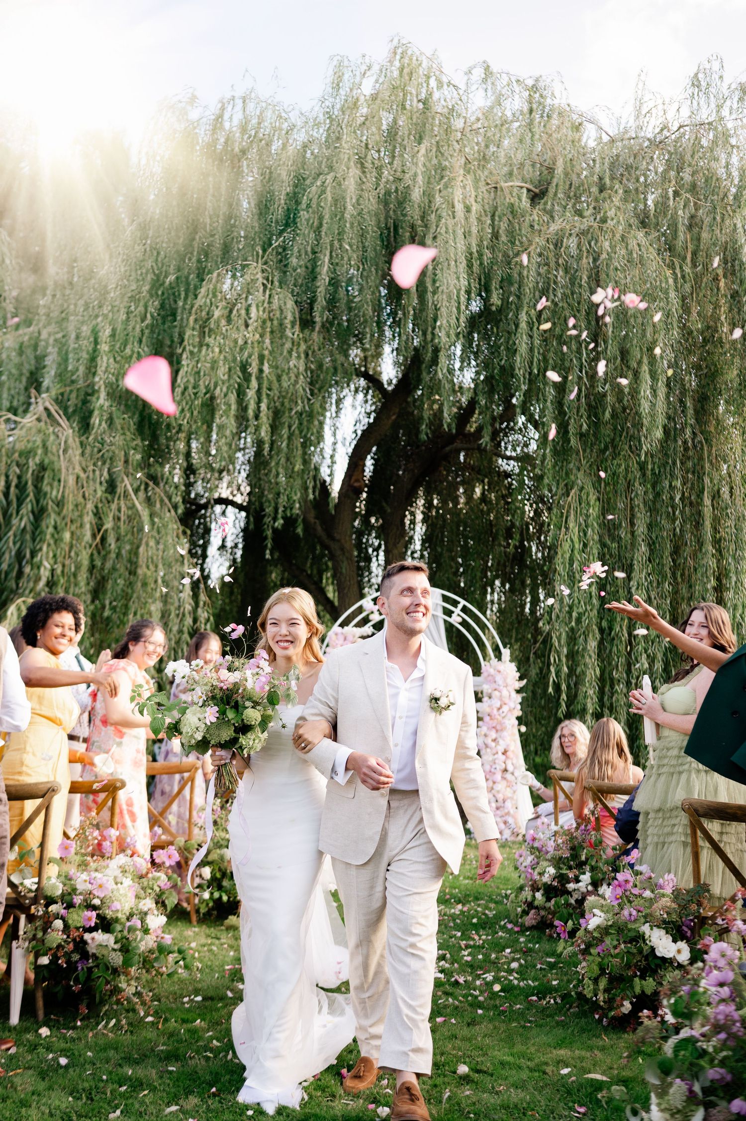 Joyful newlyweds walk down outdoor aisle as guests toss pink petals under a willow tree.