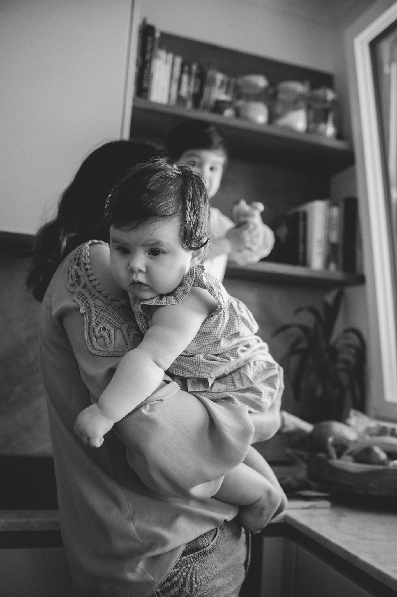 Black and white photo of a mother holding her baby in a kitchen while another child plays in the background.