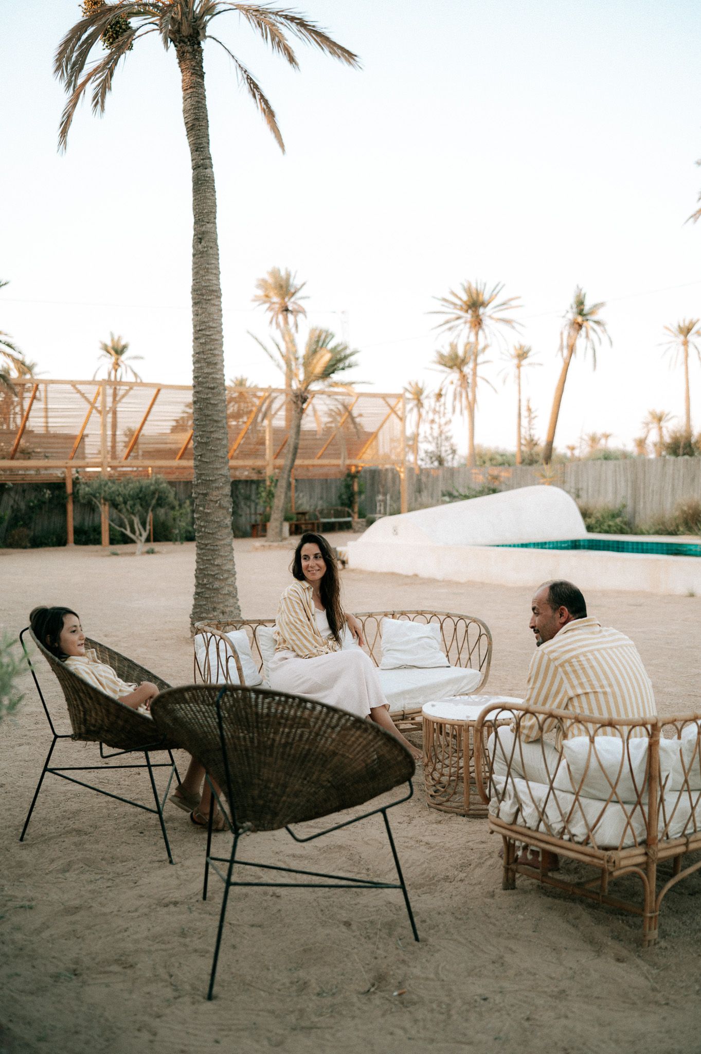Three people relaxing on rattan chairs in sandy outdoor lounge area surrounded by palm trees near a pool.