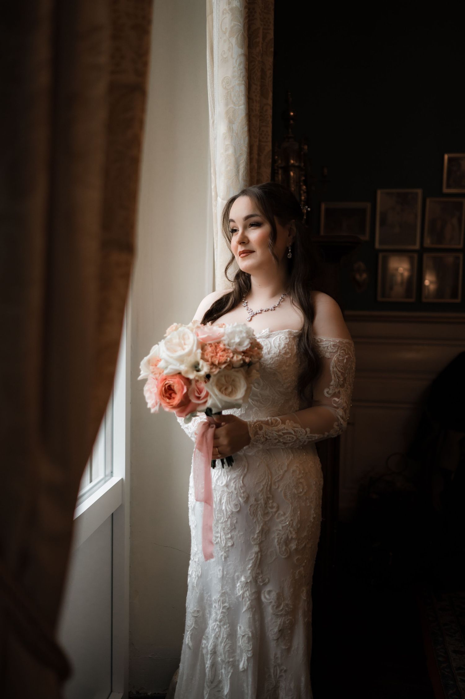 Bride in lace wedding gown holding a pink and white bouquet while gazing out a tall window.