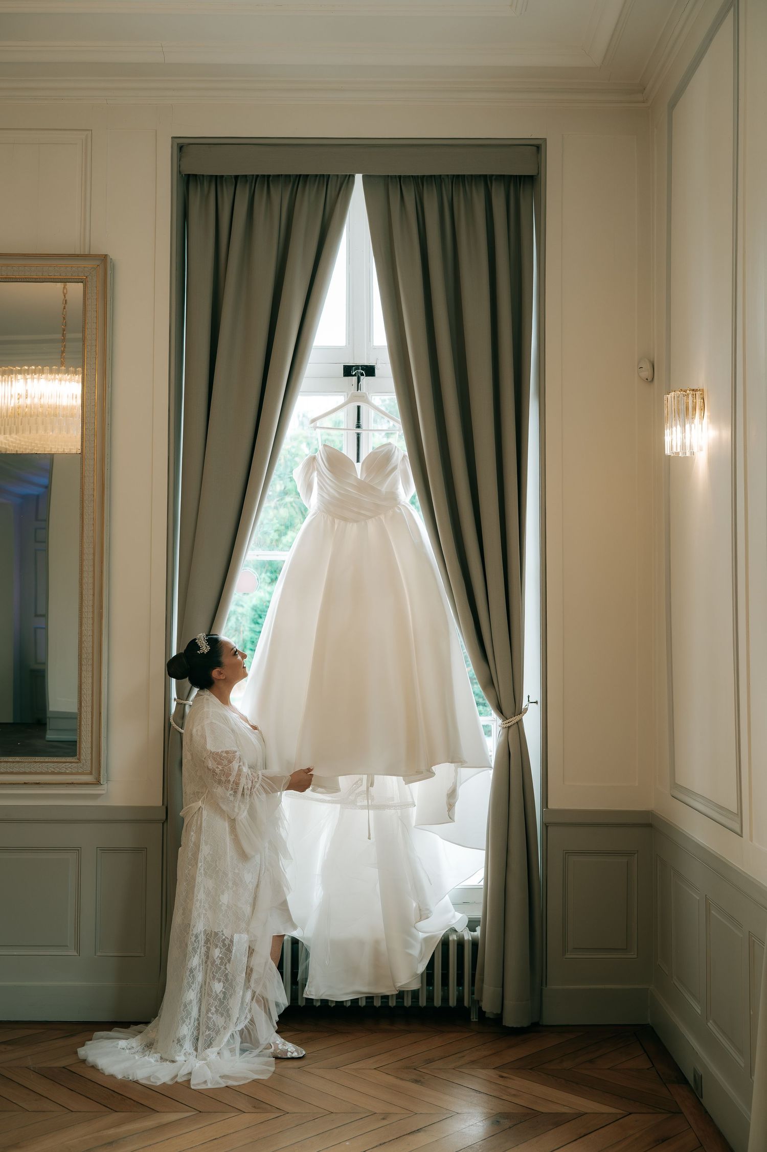 Bride in lace robe admiring her elegant white ball gown hanging in a window of a classic French château.