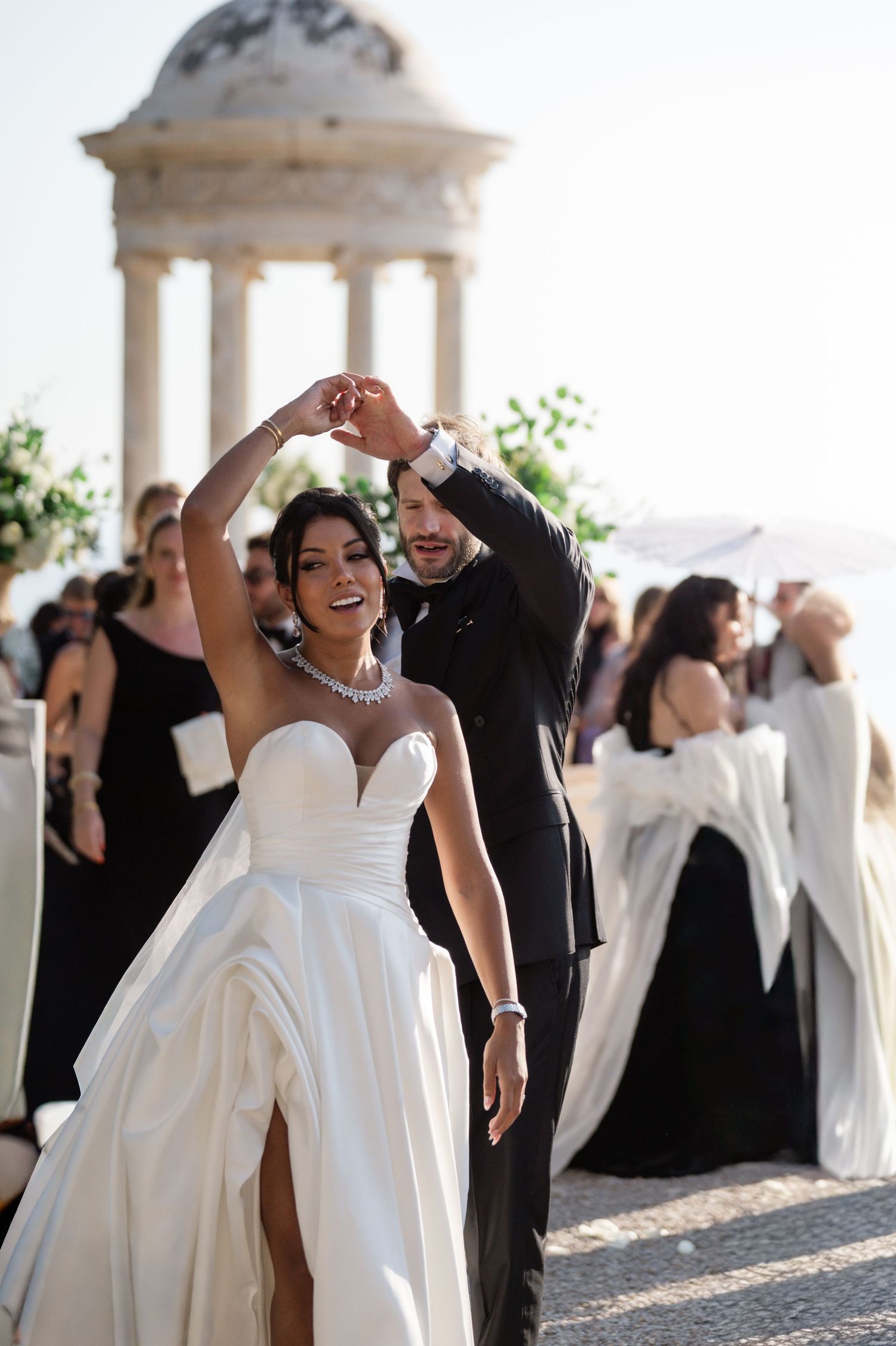 Jubilant newlyweds celebrate after their ceremony near a classical rotunda, bride raising her arm joyfully.