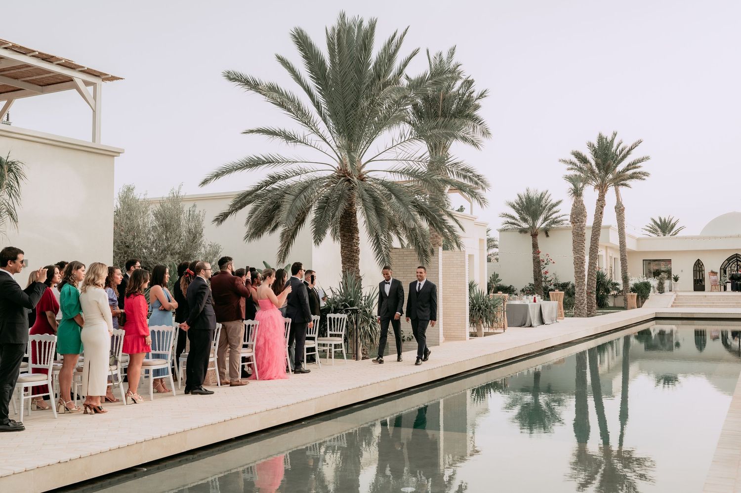Wedding ceremony guests gathered beside a serene reflecting pool surrounded by palm trees at an outdoor venue.