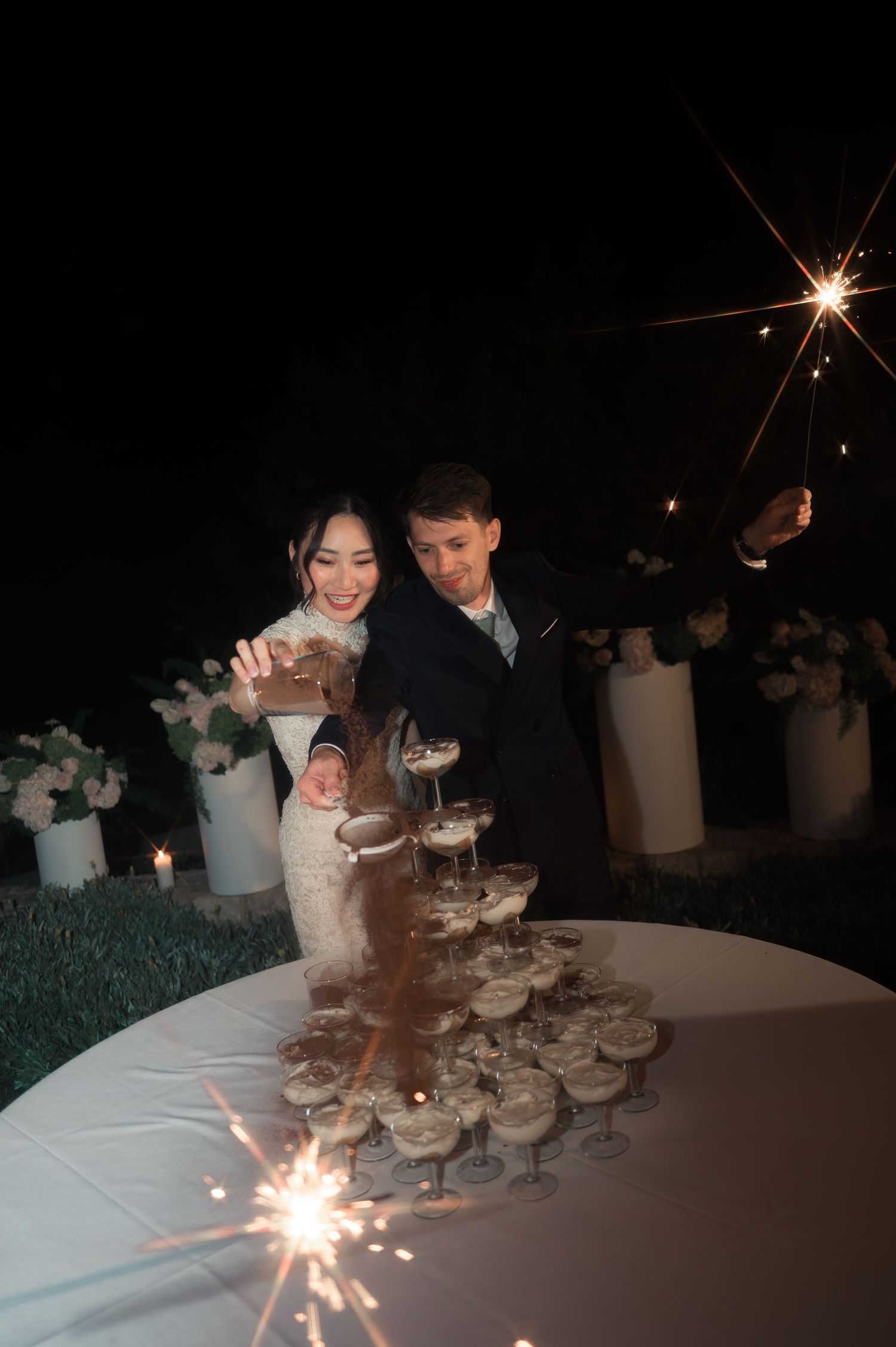 Bride and groom pouring champagne into a tower of glasses while sparklers light the night.