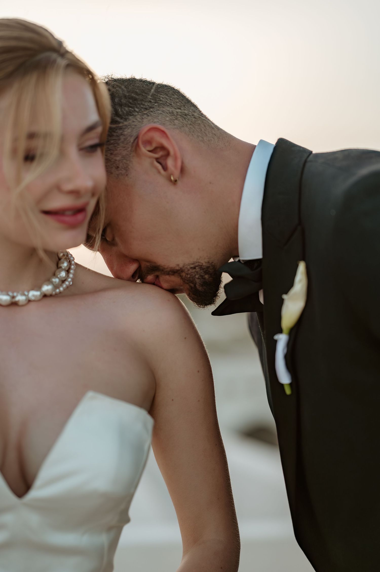 Romantic wedding moment as groom in black tuxedo gently kisses brides shoulder at a beach sunset ceremony.