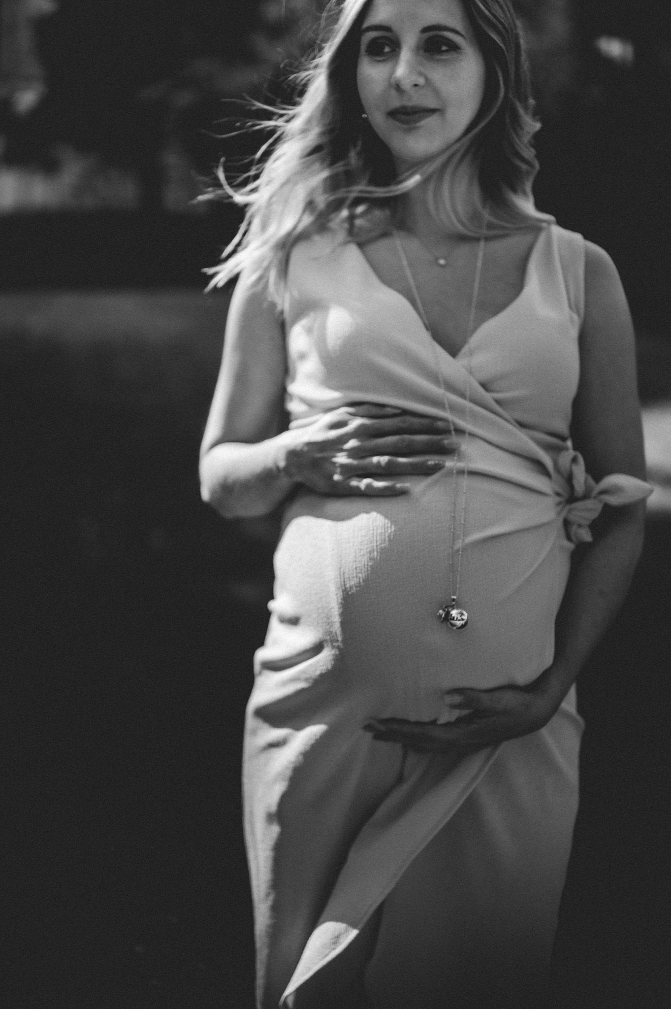 Black and white photo of a pregnant woman in a sleeveless dress cradling her baby bump outdoors.