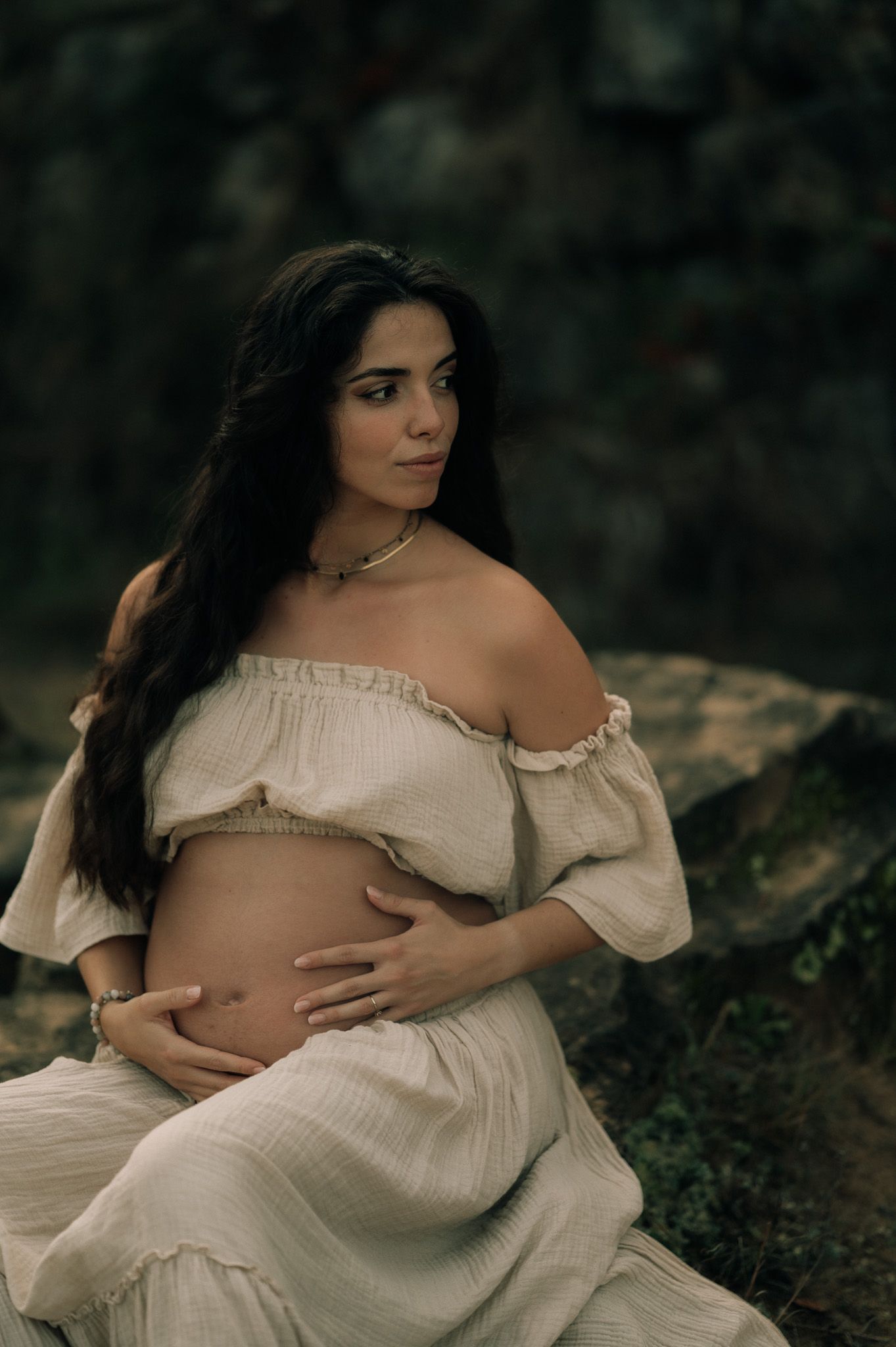 Pregnant woman in white off-shoulder dress cradling baby bump outdoors against stone wall backdrop.