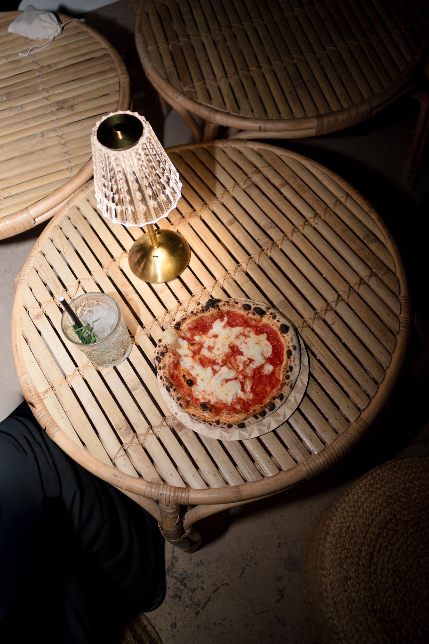 Margherita pizza and cocktail on a round rattan table with a small gold lamp.