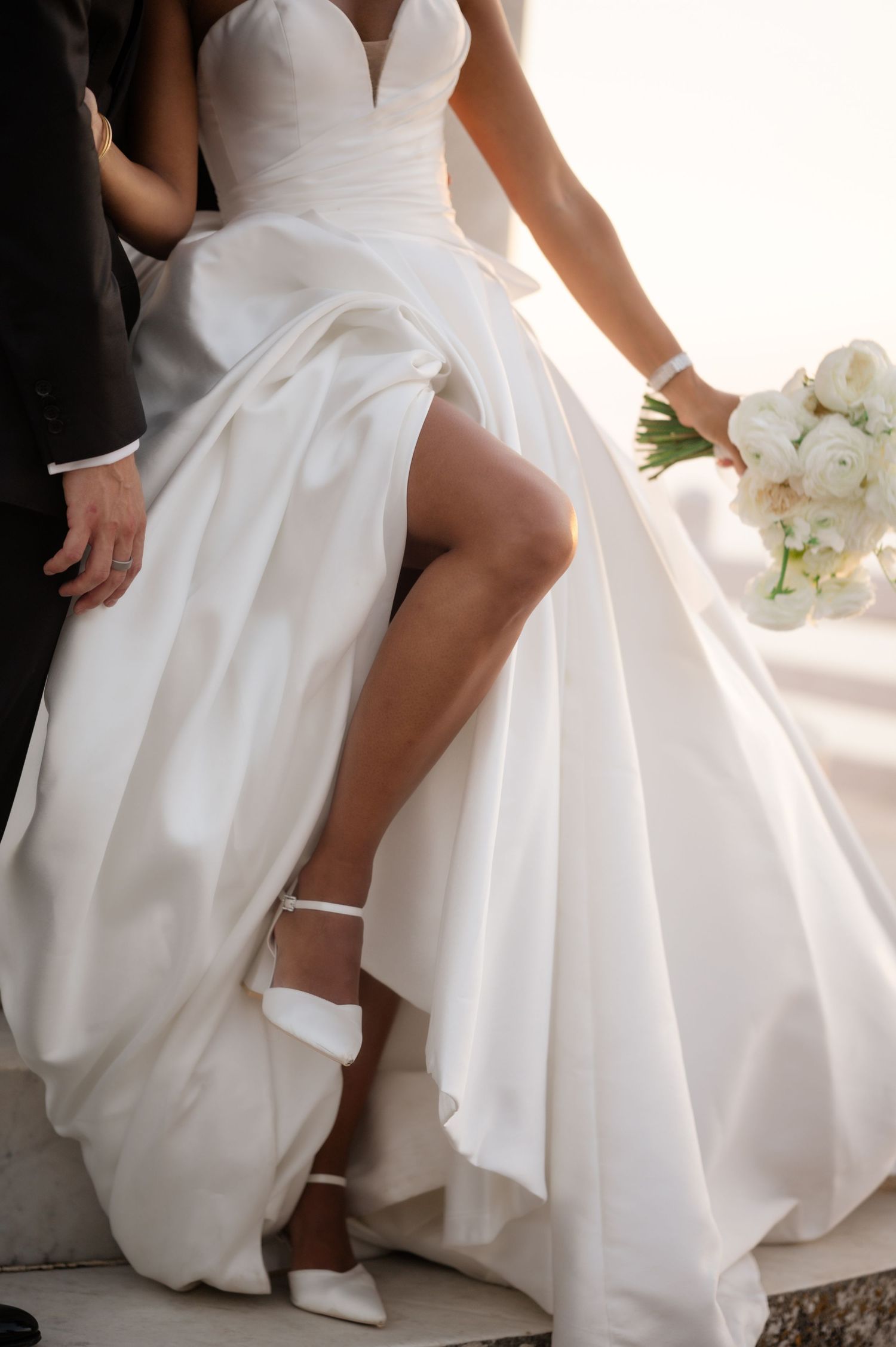 Bride in elegant white satin ball gown with high slit and white ankle strap heels holding white floral bouquet.