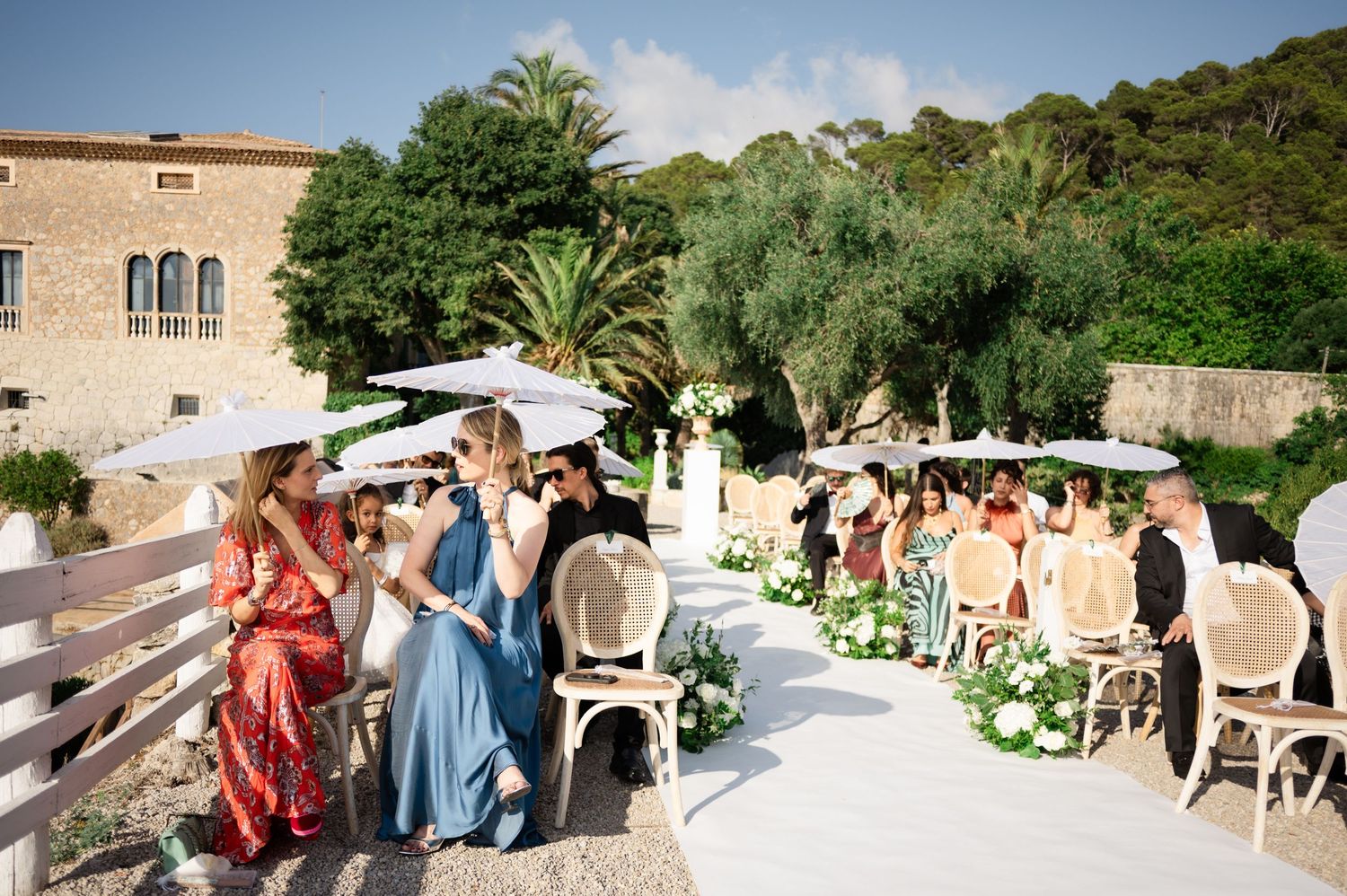 Guests with white parasols seated at an outdoor wedding ceremony in a Mediterranean garden.