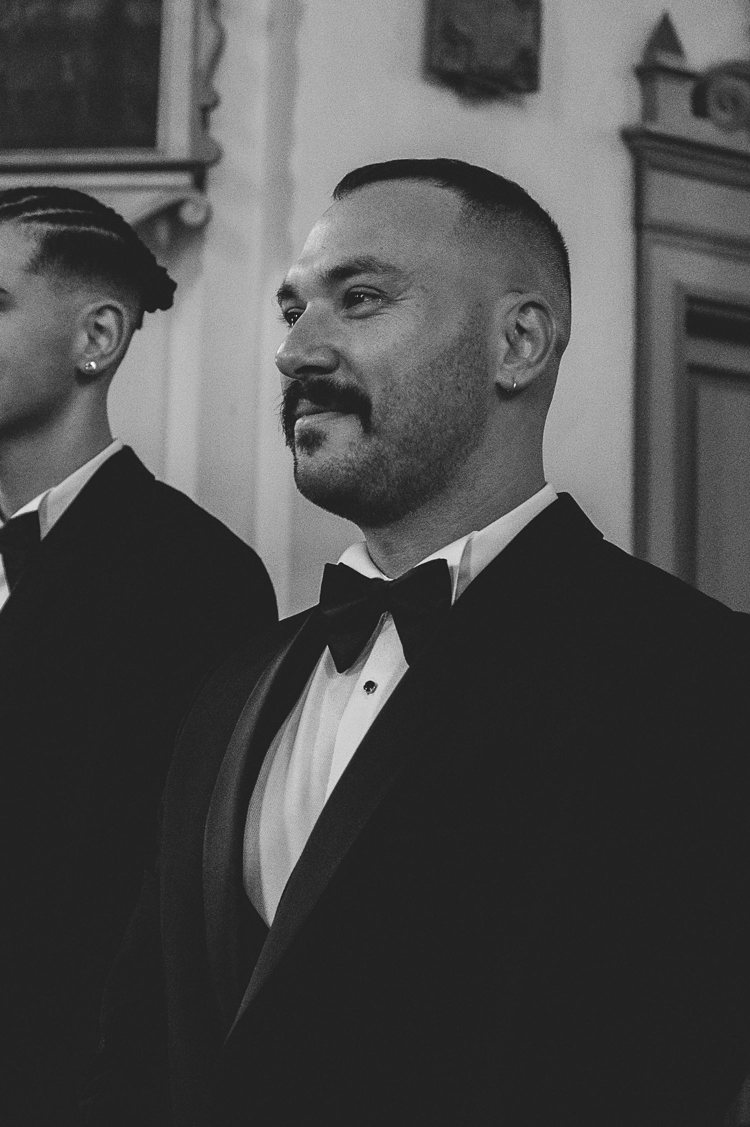 Black and white portrait of a groom in a tuxedo with bow tie and mustache standing at a wedding ceremony.