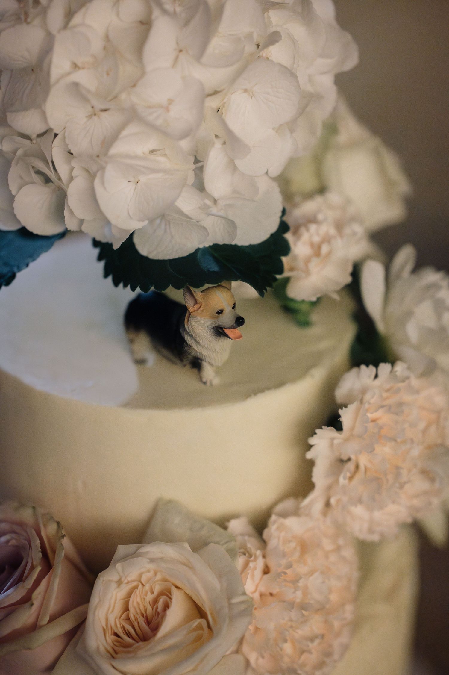 Close-up of wedding cake detail featuring a corgi figurine nestled among white hydrangeas and pink roses.