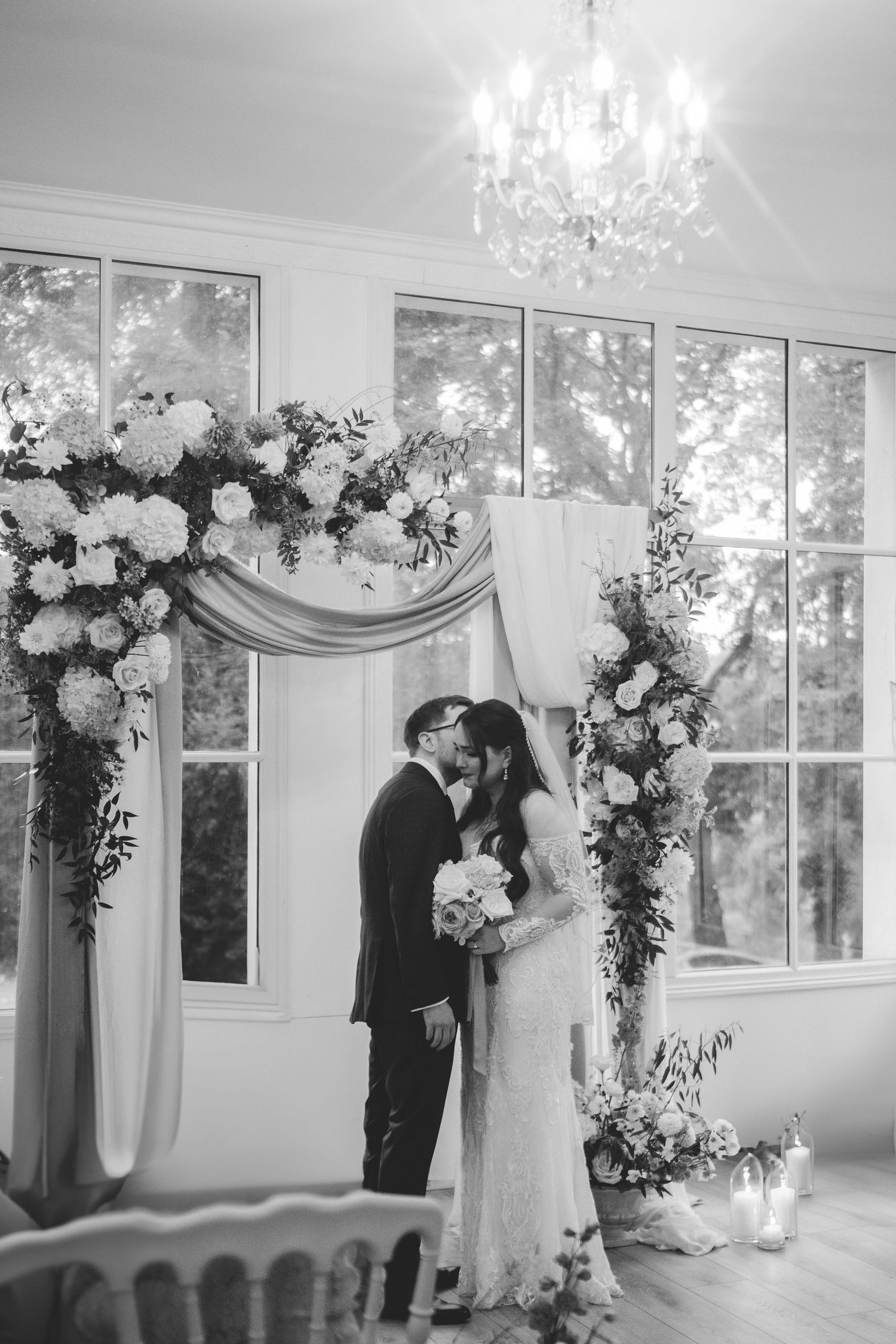 Black and white photo of bride and groom sharing first kiss under a chandelier and floral arch.