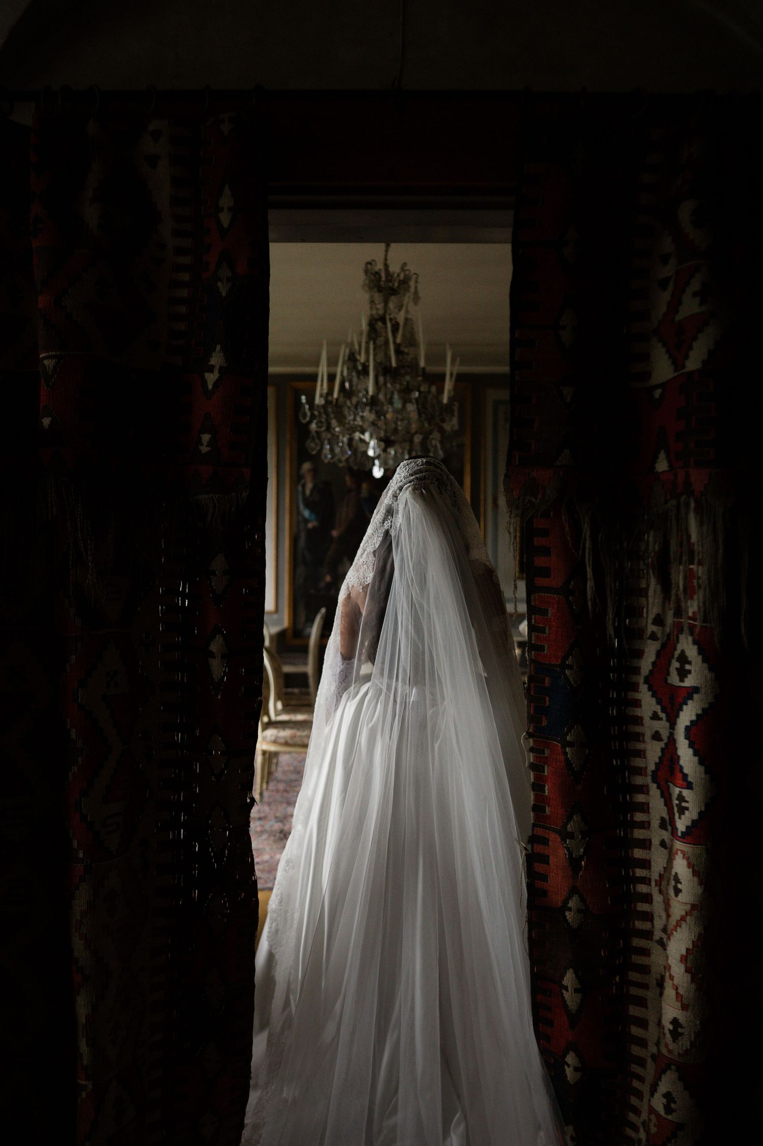 Mysterious bride in white veil seen through doorway framed by colorful traditional woven carpets.
