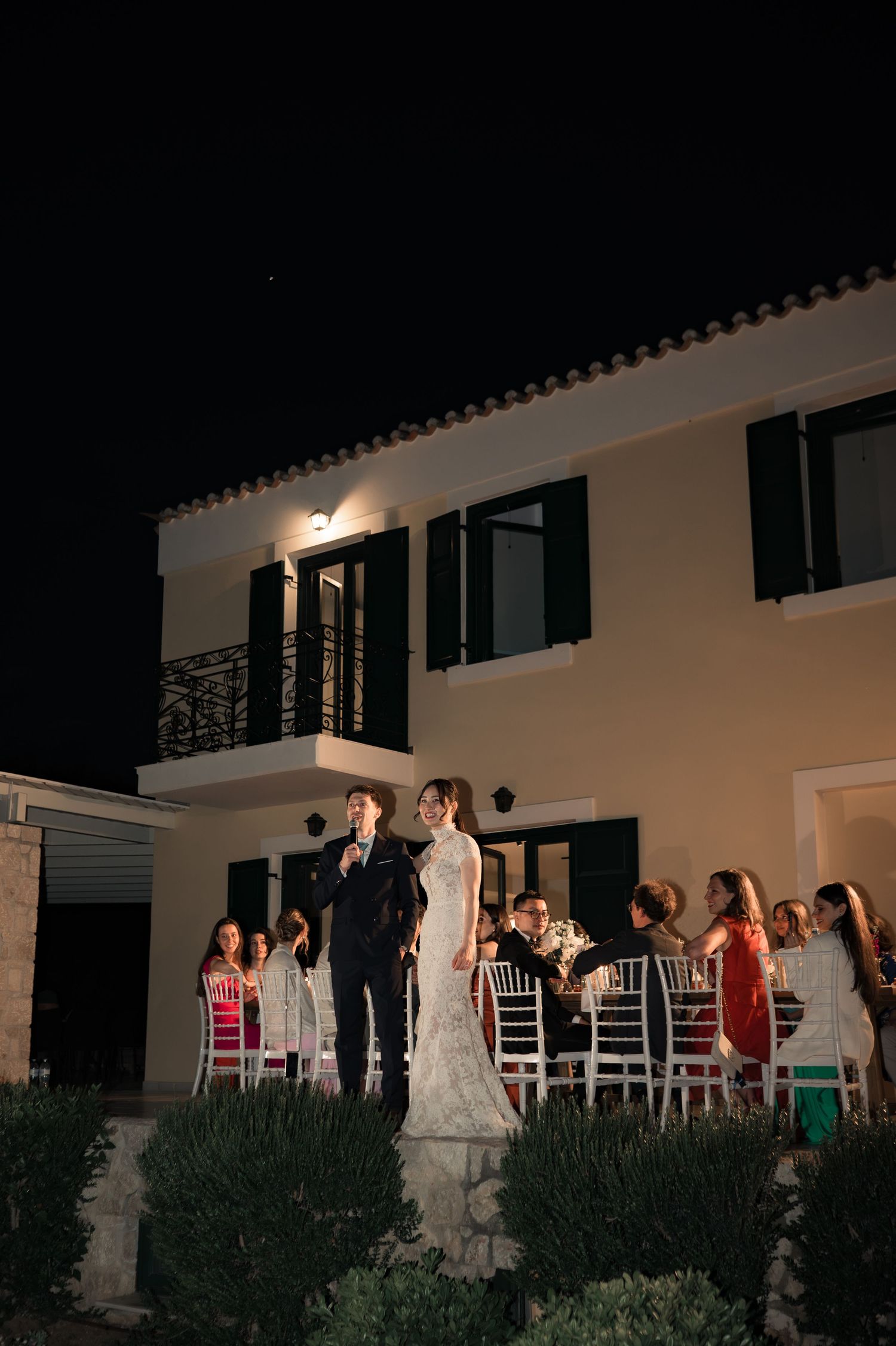 Bride and groom stand together outside an illuminated villa during their evening wedding reception.