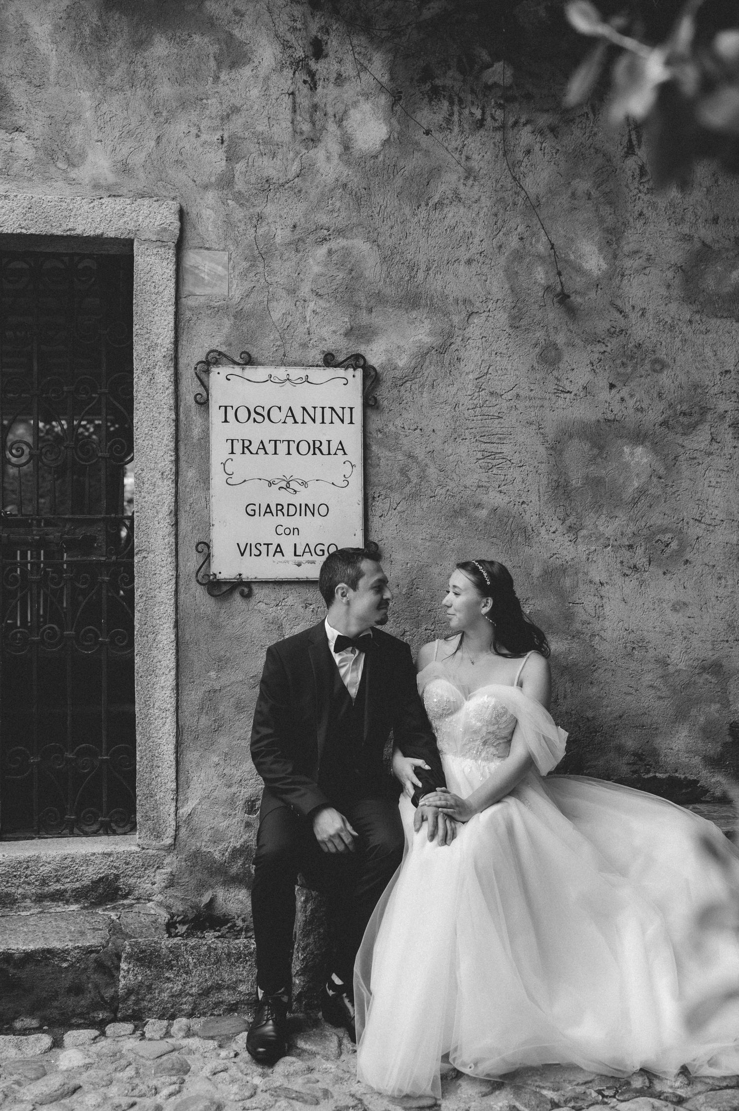 Black and white photo of bride and groom posing by a rustic trattoria wall in Italy.