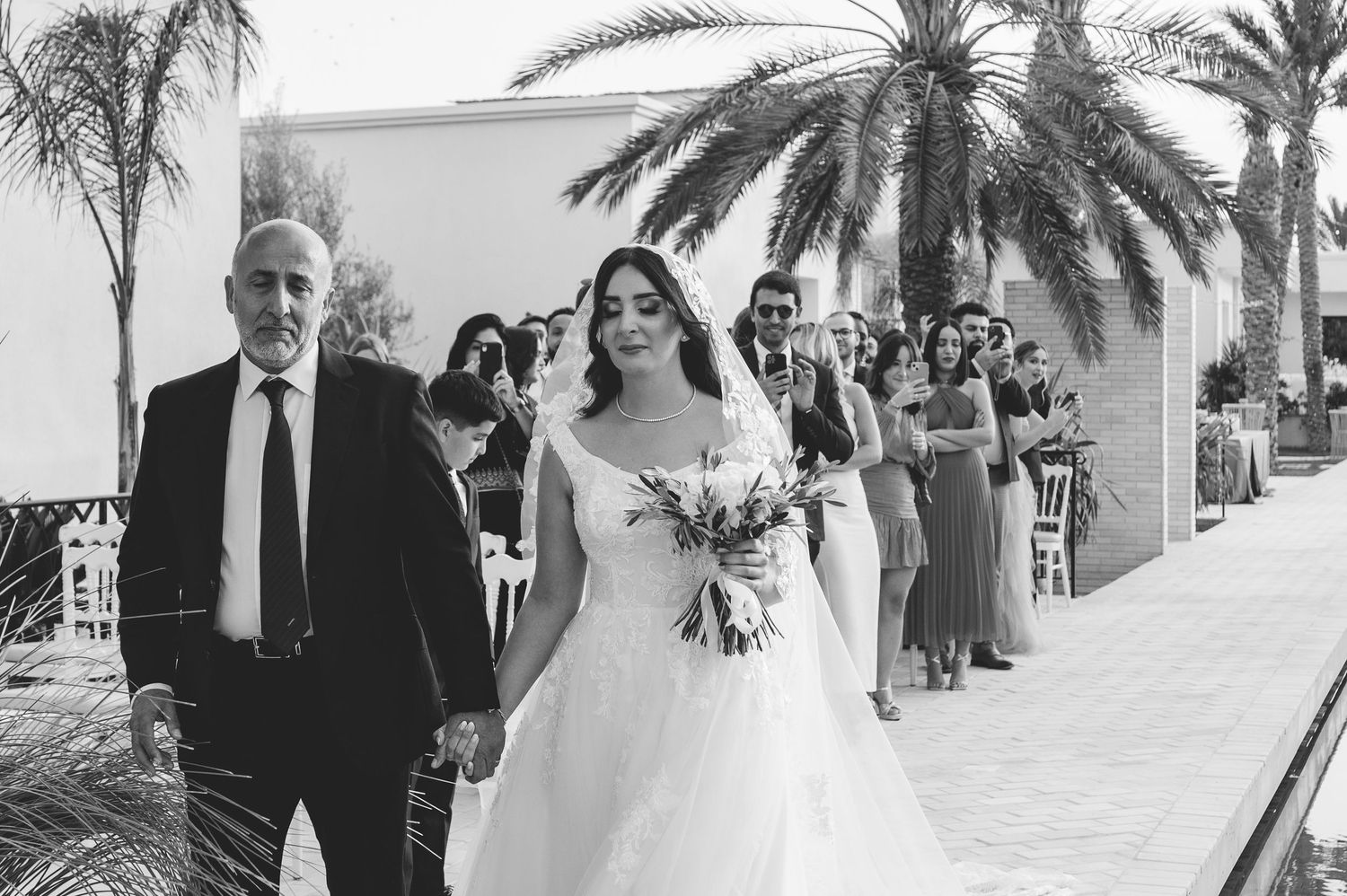 Father walking bride down the aisle in a white wedding gown past seated guests at an outdoor ceremony.
