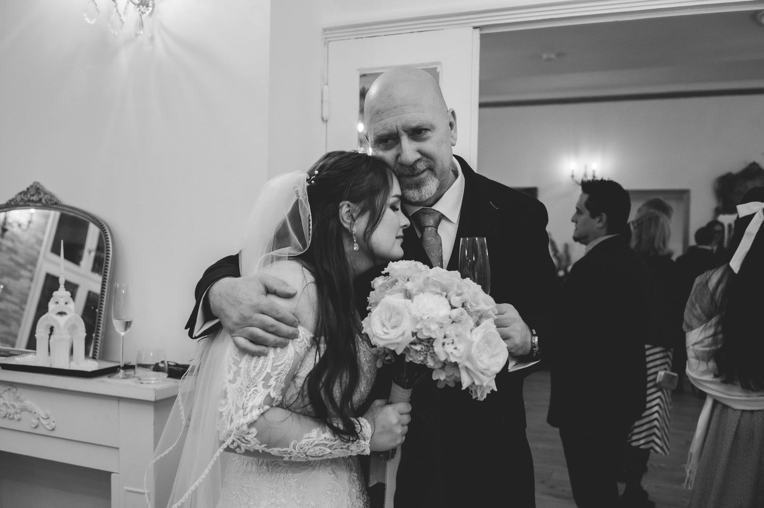 Black and white photo of bride embracing older man holding bouquet at wedding reception.