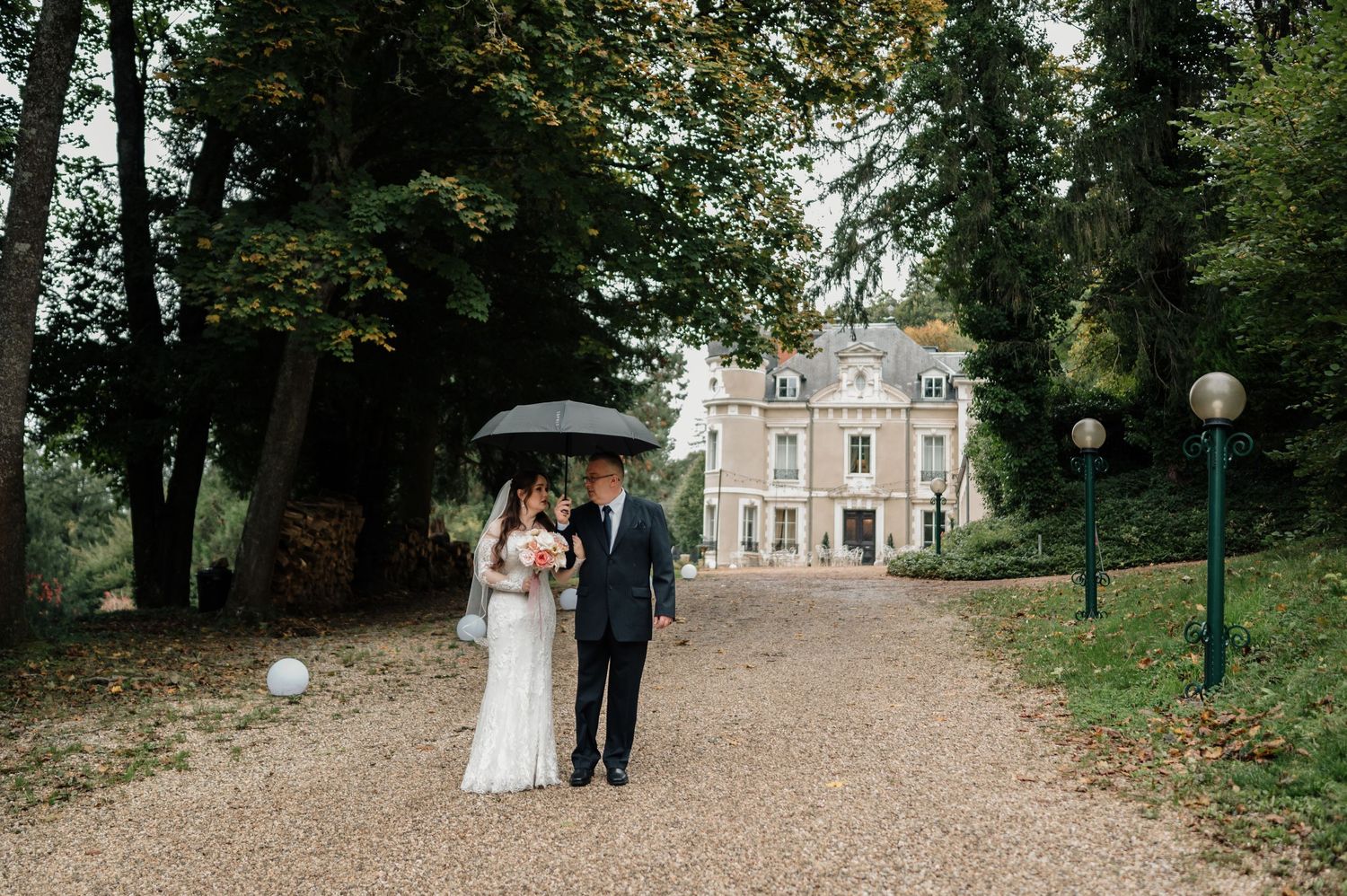 Bride and escort walk umbrella-shaded gravel path toward a grand chateau on a rainy wedding day.